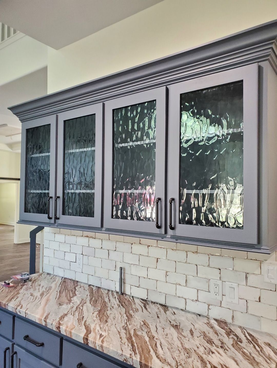 Dark gray upper cabinets with glass panels above a beige brick backsplash and marble countertop in a kitchen.
