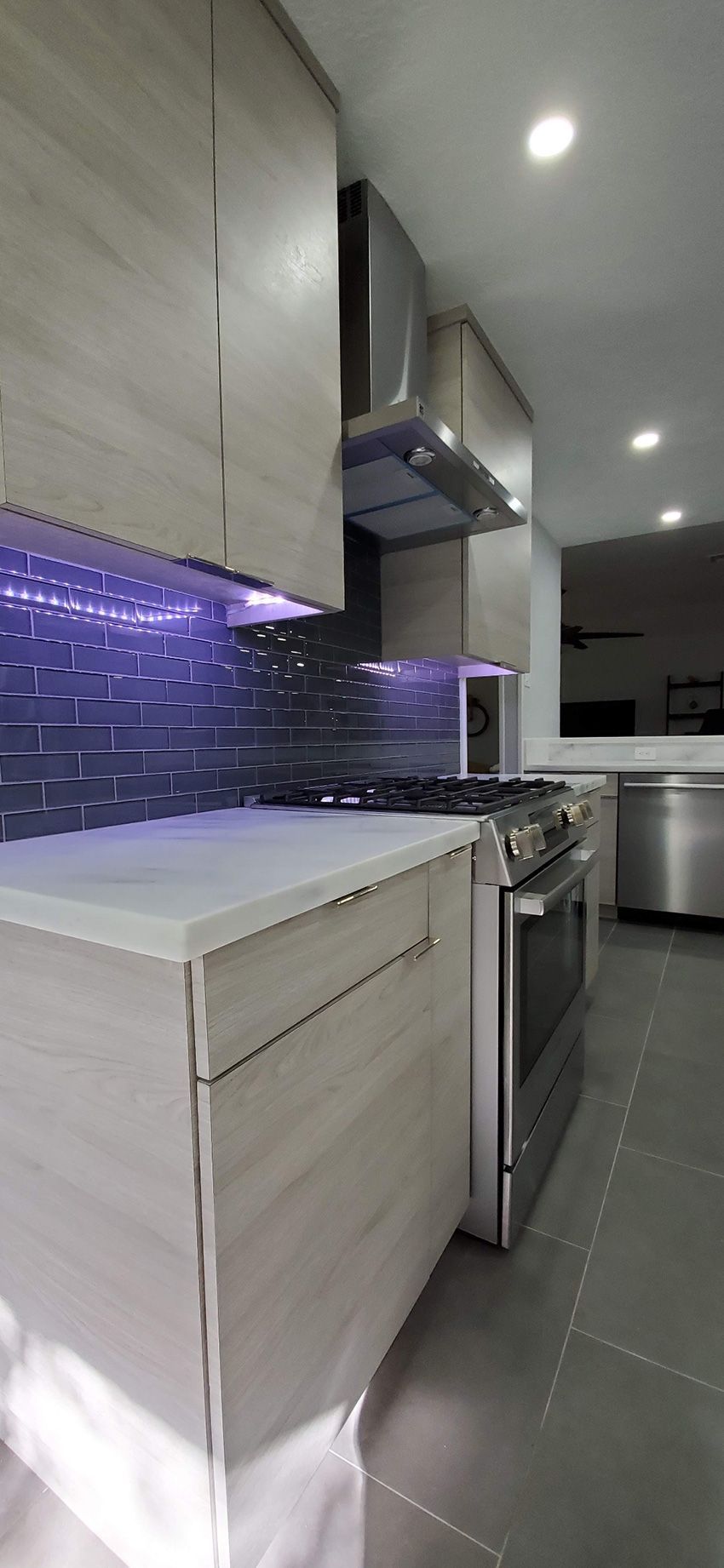 Modern kitchen with light wood cabinets, white countertops, dark gray tiled backsplash with purple LED lights, and stove.