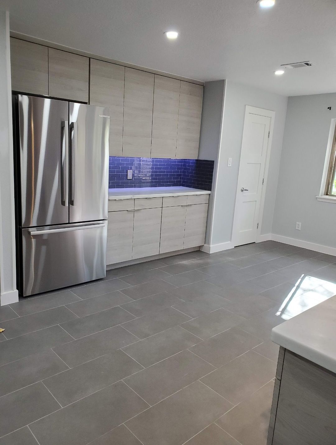 A modern kitchen featuring light wood cabinetry, a stainless steel refrigerator, blue backsplash, and grey tile floors.