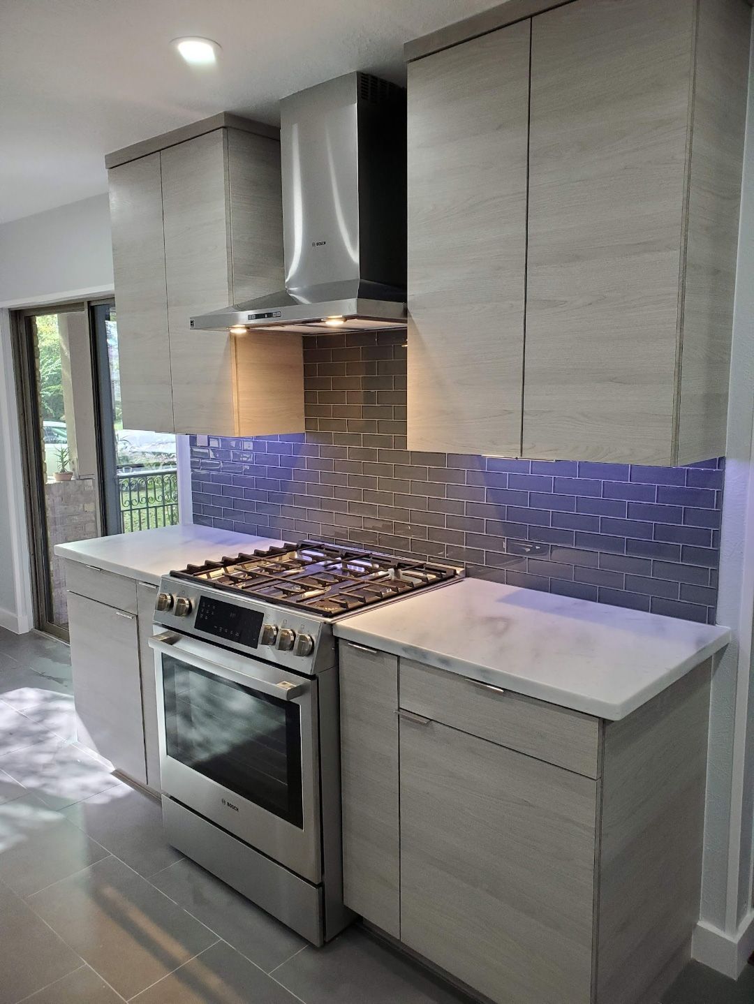 A modern kitchen featuring light wood grain cabinets, a stainless steel gas range, a vent hood, and blue tiled backsplash.