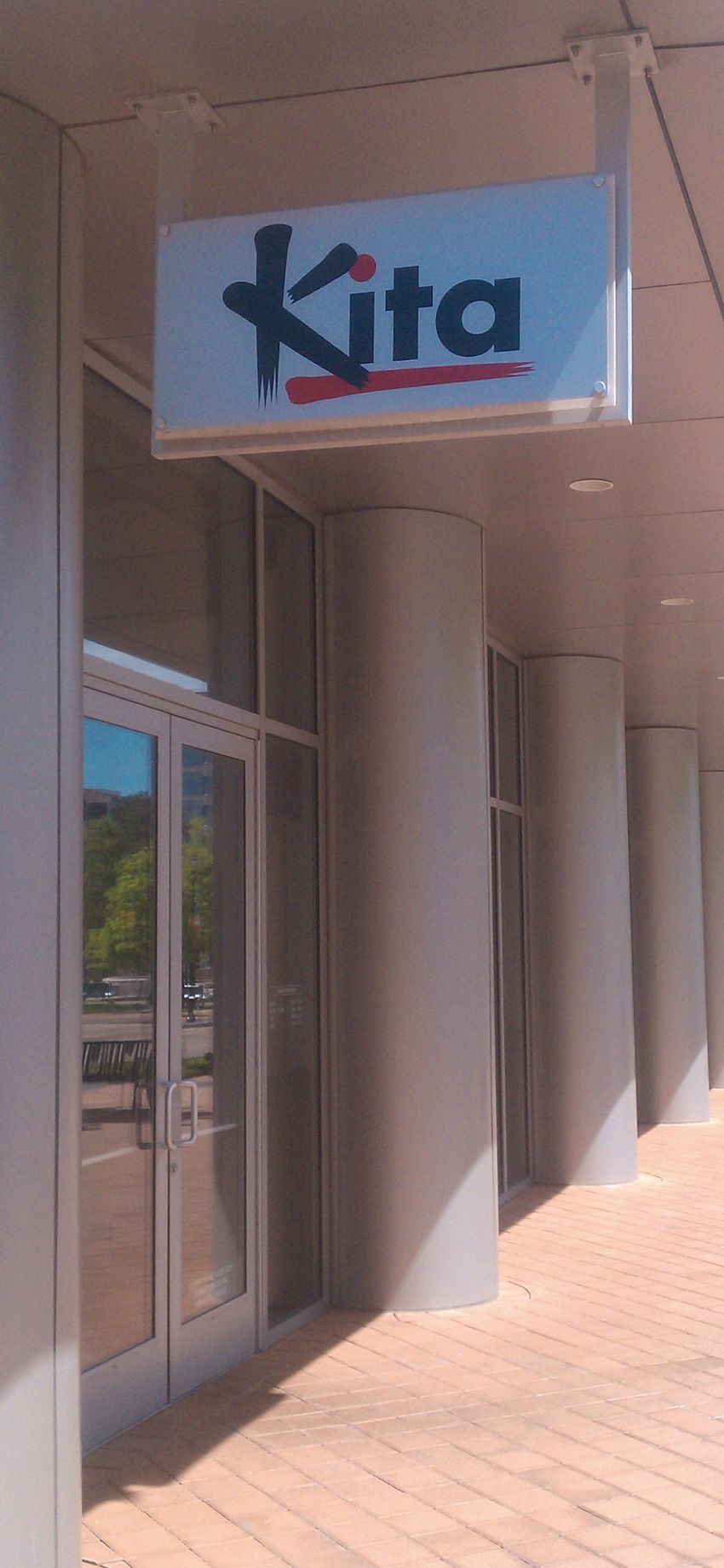 An eye-level shot of a building exterior with a Kita sign hanging above a glass entrance door near white pillars.