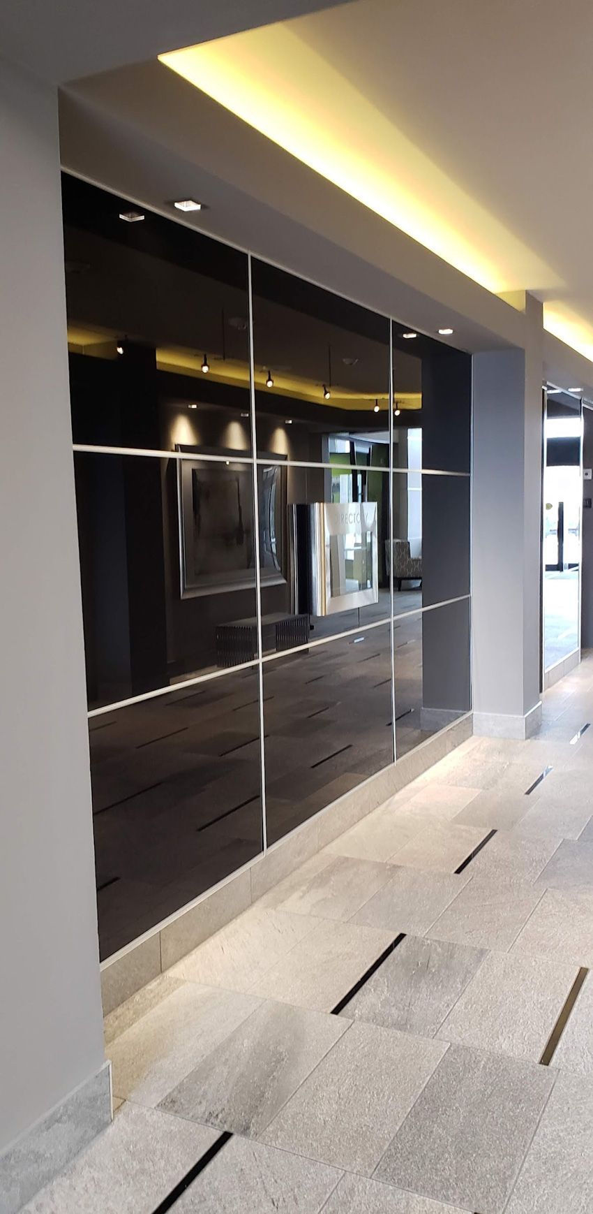 A dark reflective glass wall in a hallway with a light-colored tiled floor featuring embedded dark linear accents.