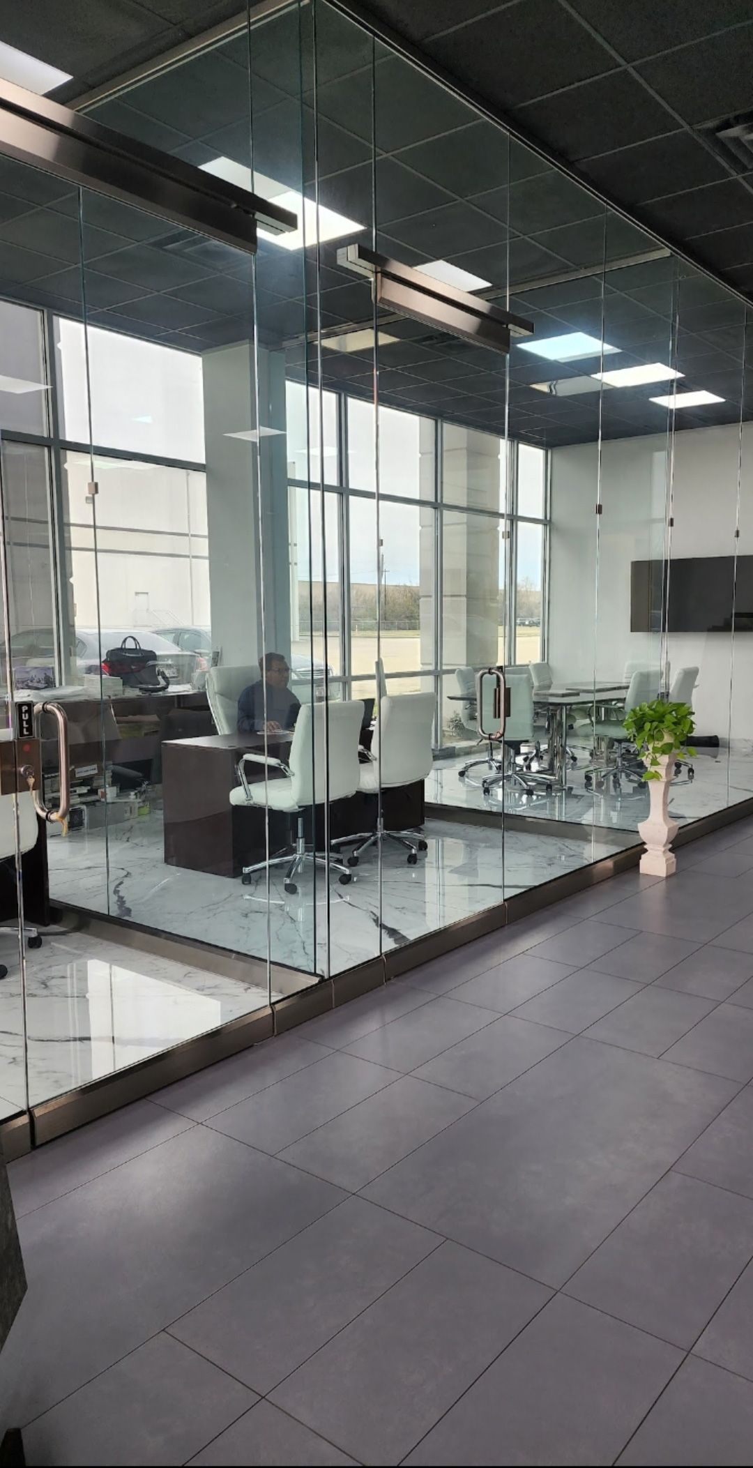 A modern office features a glass-walled conference room with white chairs, dark desks, and floor-to-ceiling windows.