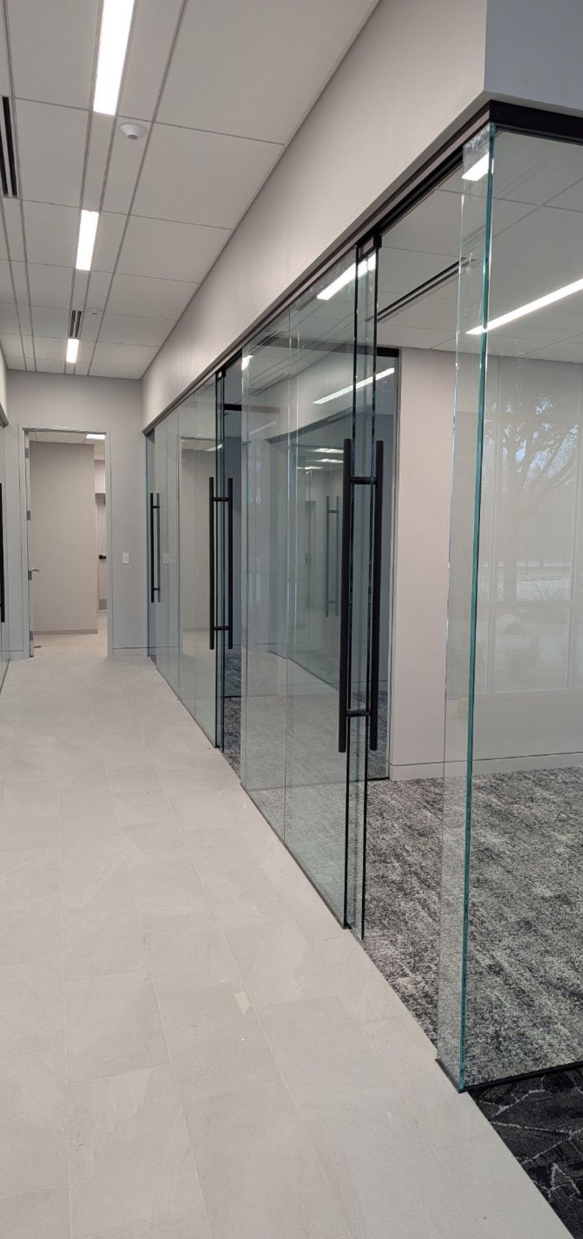 A modern office hallway with a tiled floor, featuring glass-walled conference rooms with black handles and metal frames.