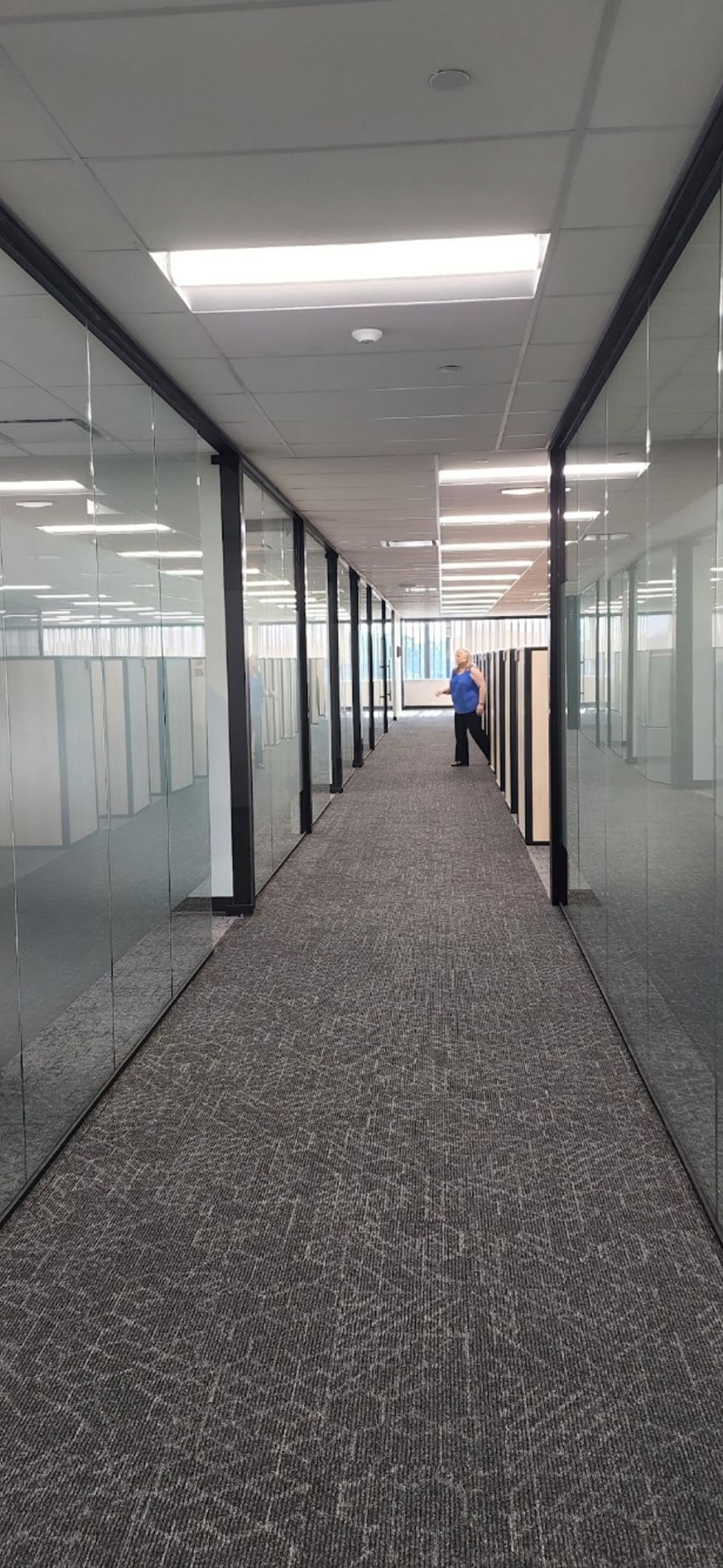 A person walks down a long, narrow office hallway lined with glass-walled rooms and grey speckled carpeting.