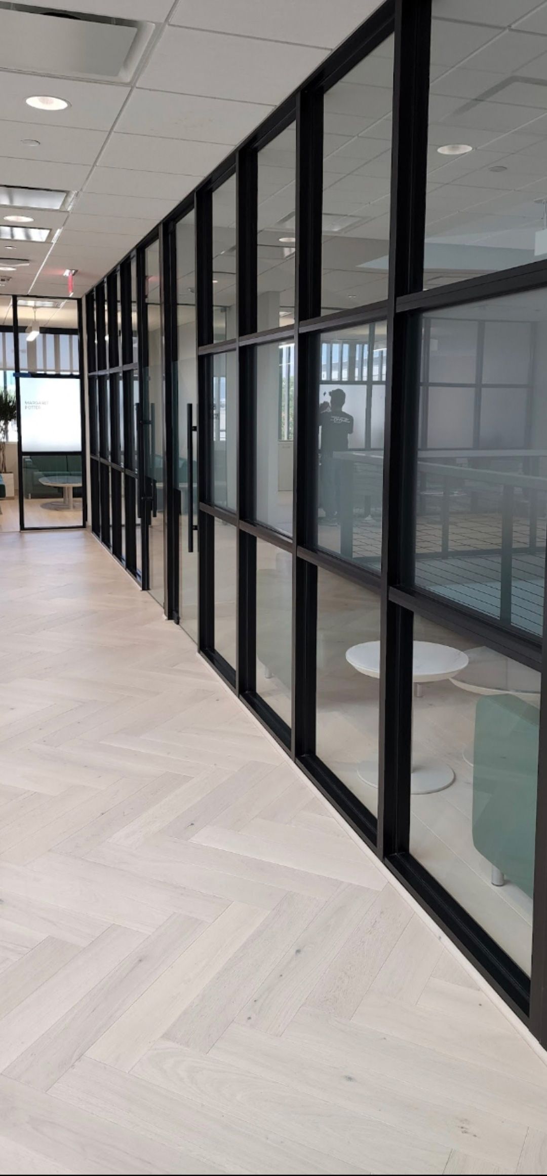 A view down a bright office hallway with light wood floors and a wall of black-framed glass partitions.