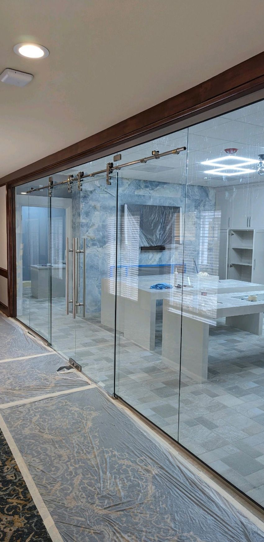 A sliding glass partition wall separates a corridor from an office space with white desks and a marble accent wall.