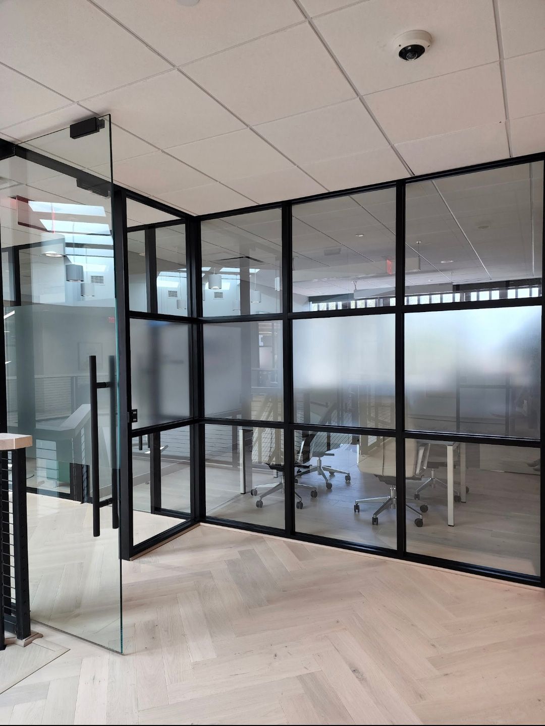 Office interior featuring a glass-partitioned meeting room with black frames and frosted glass panels on light wood floors.