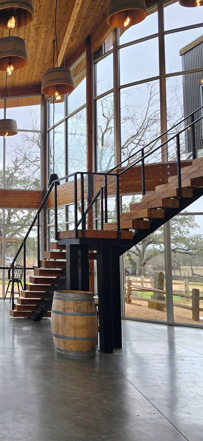 Modern interior featuring a wooden staircase, black metal railings, a wooden barrel, and pendant lights by a large window.