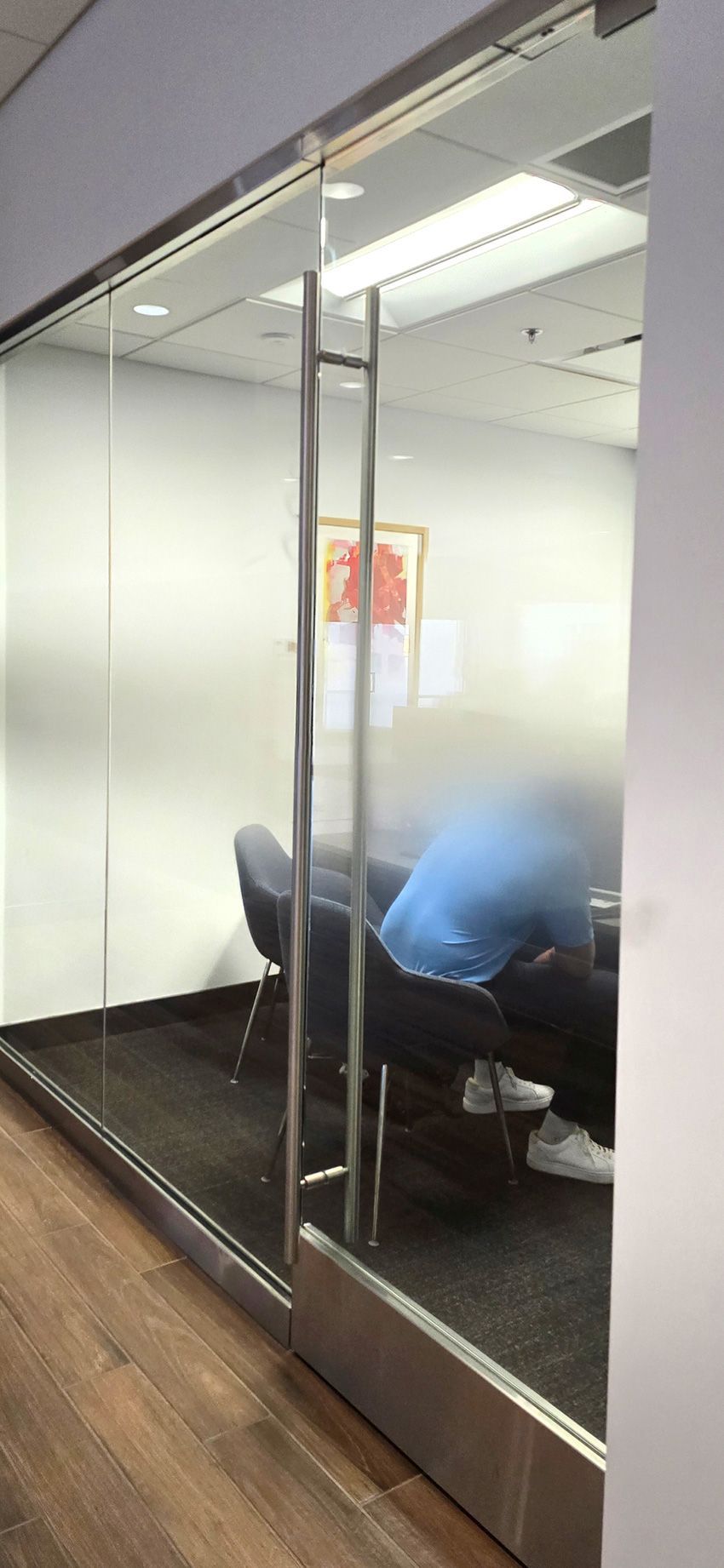A person wearing a blue shirt sits in a glass-walled conference room with dark flooring.