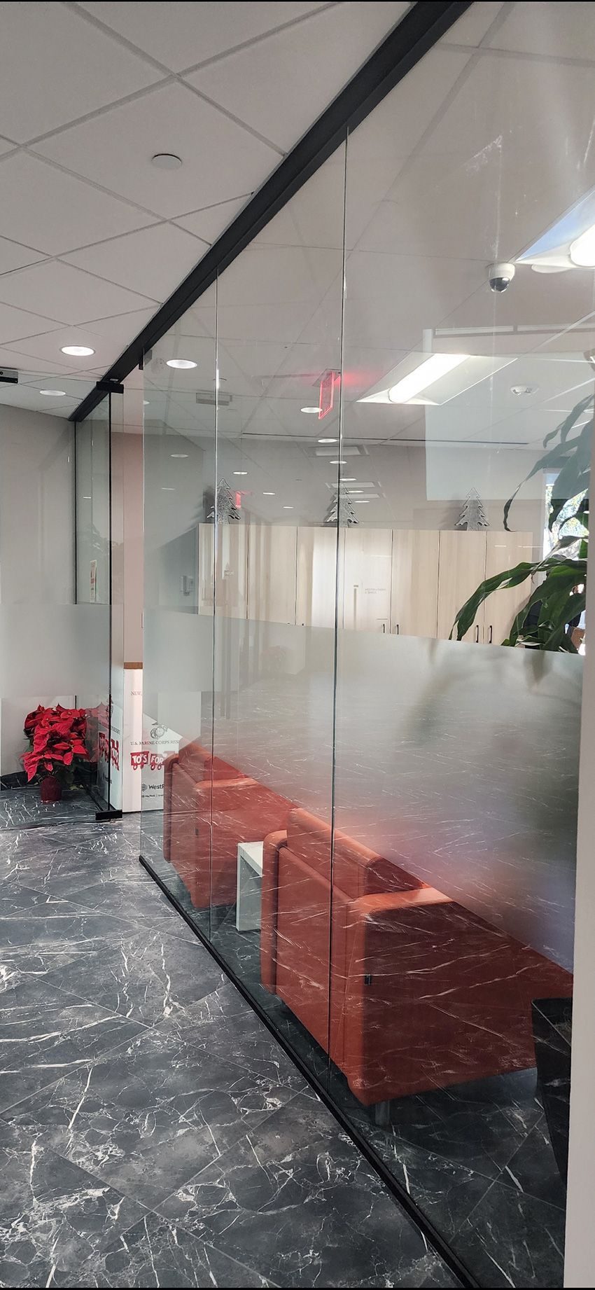 Two red armchairs sit behind a glass partition in a bright, modern office with gray marble flooring.