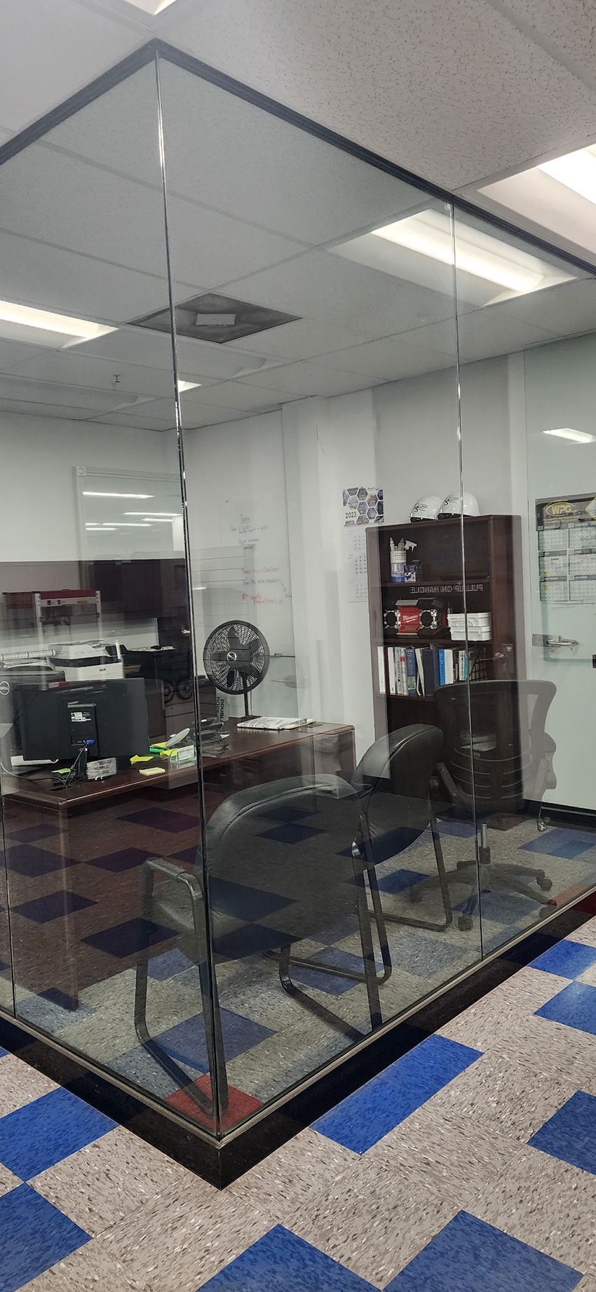 A modern glass-walled office cubicle featuring a black desk, computer, swivel chair, and shelving against a blue-tiled floor.