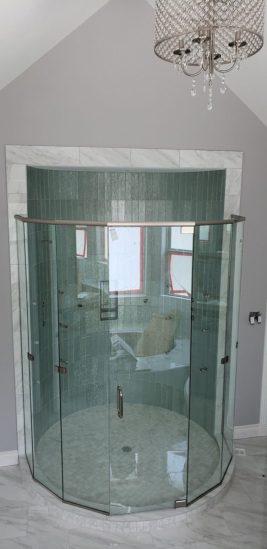 A modern, curved glass walk-in shower with chrome hardware, set against a grey wall with a decorative ceiling chandelier.
