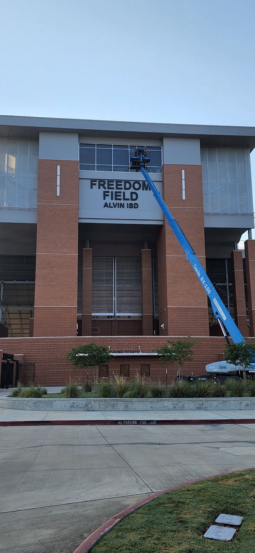 A blue boom lift extends toward the sign for Freedom Field on the brick facade of a building under construction.