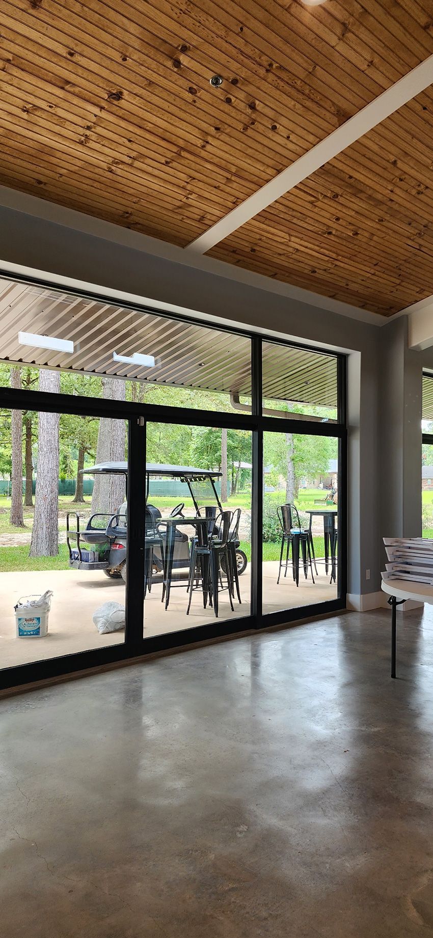 A modern, spacious room with large glass doors looking out onto a patio with a golf cart and trees under a wood ceiling.