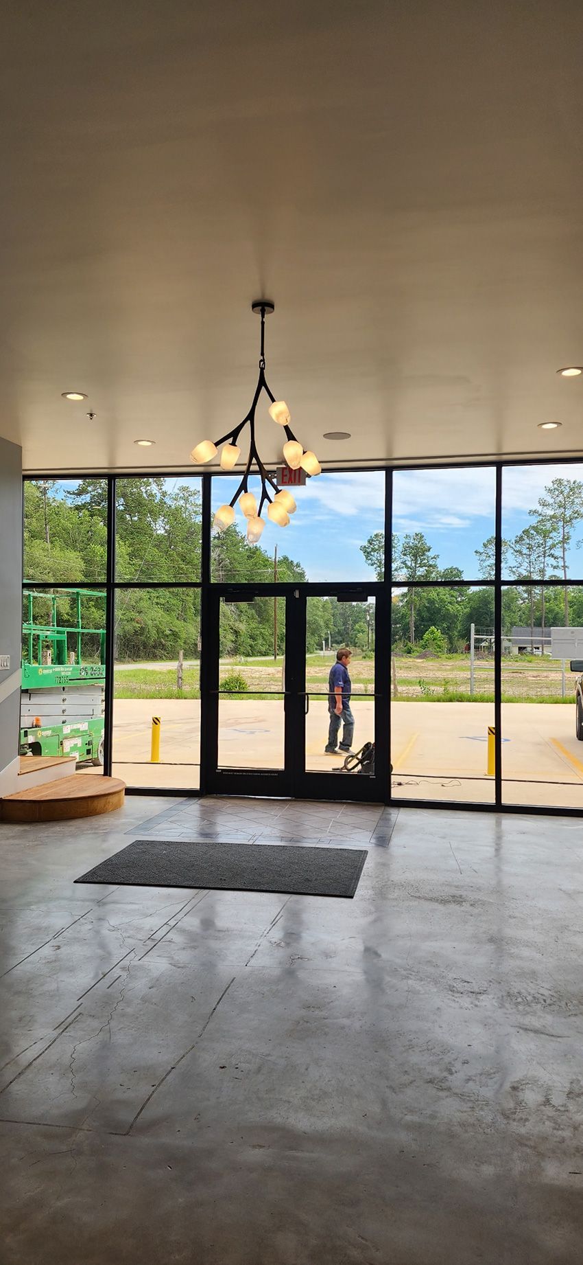 Modern building lobby with concrete floors, a branch-style chandelier, and large glass windows looking out to a parking lot.