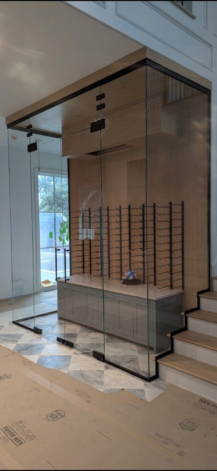 A modern display area with a glass-enclosed wooden wall, shelving for glasses, a base cabinet, and stairs in a room.