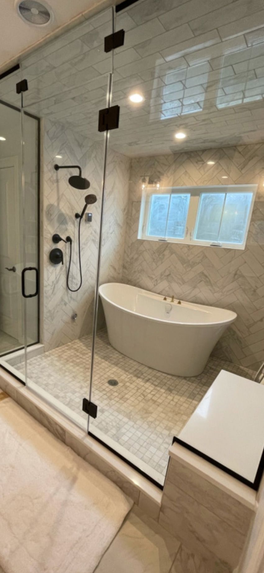 A modern walk-in shower with a glass enclosure featuring a freestanding tub, matte black fixtures, and marble tile walls.