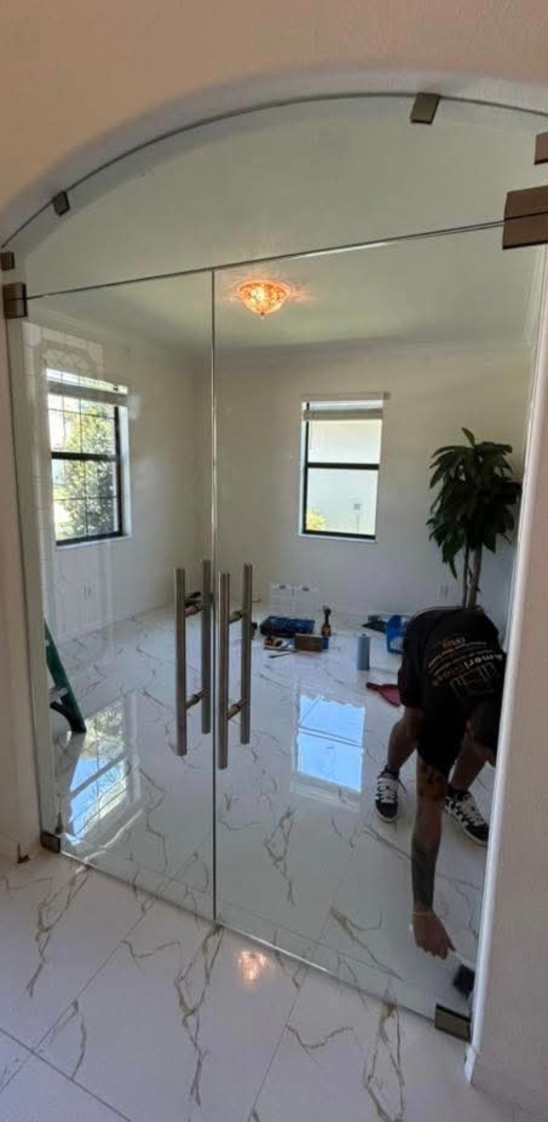 A person installing glass double doors with large metal handles in a room with white marble floors and two windows.