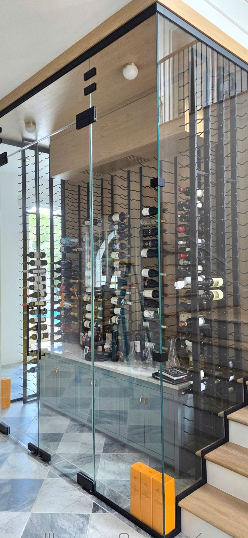 A glass-enclosed wine cellar featuring floor-to-ceiling metal racks holding wine bottles, situated next to wooden stairs.