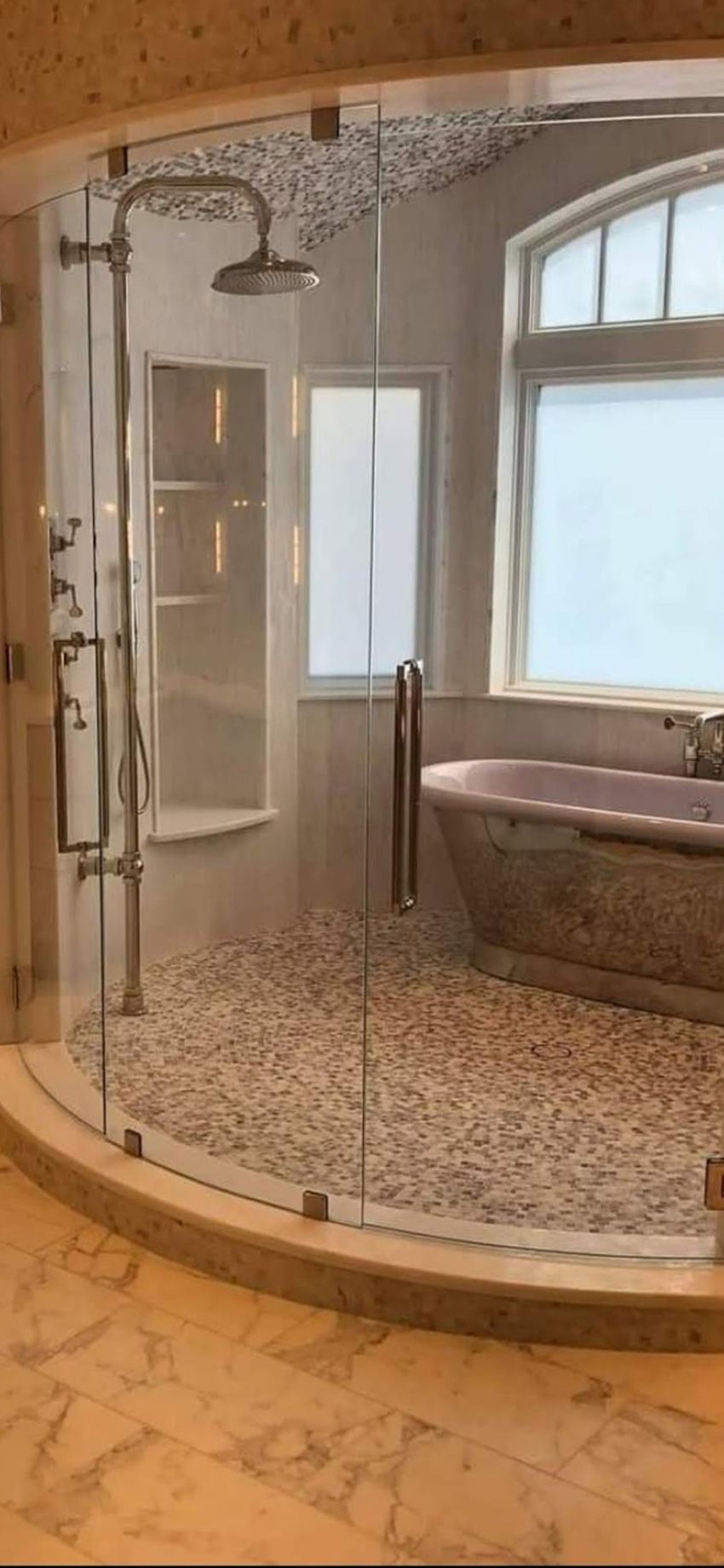 A curved glass walk-in shower featuring pebble tile flooring, a chrome rainfall showerhead, and an adjacent soaking tub.