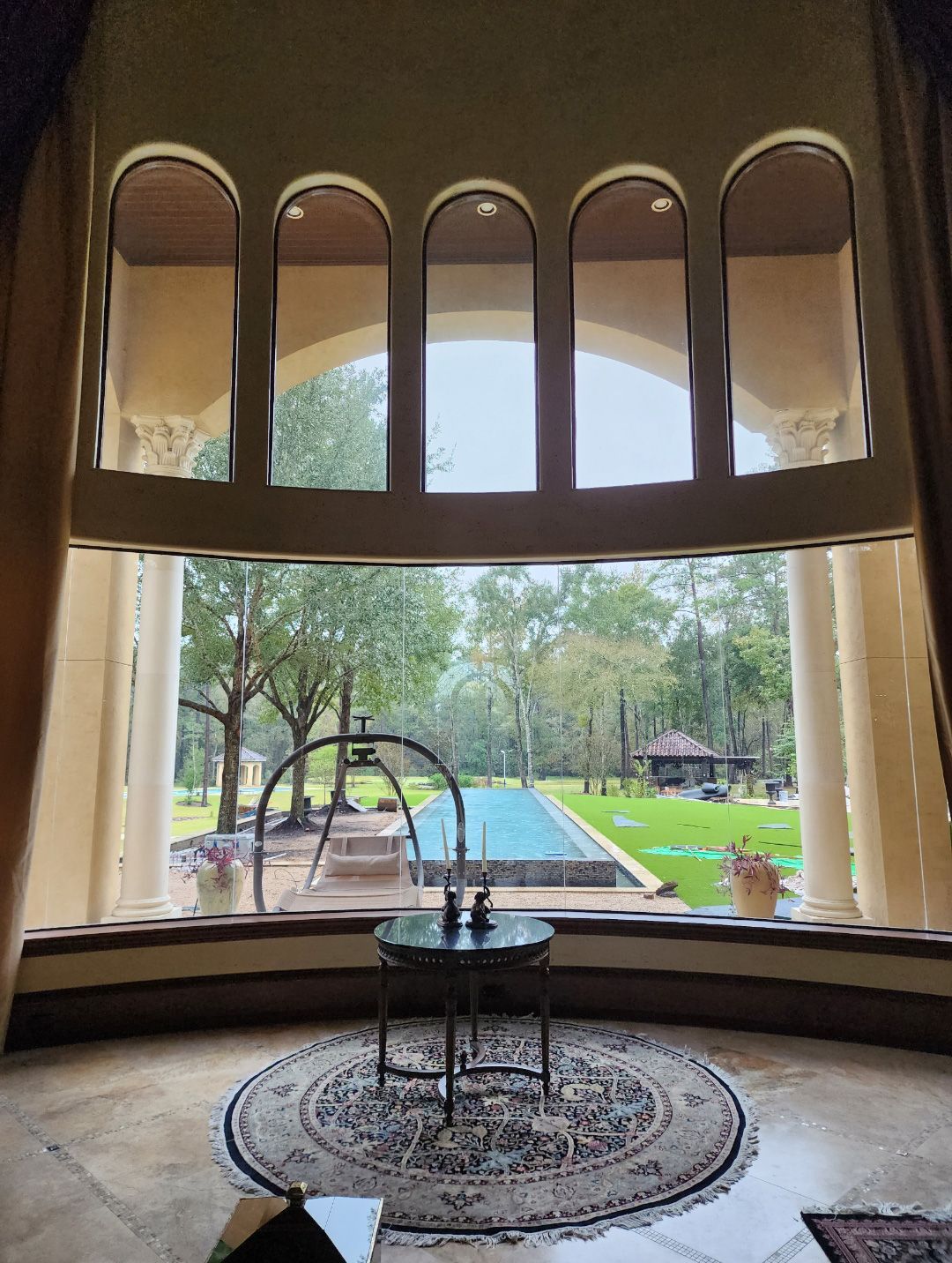 A view through a grand arched window looking out onto a landscaped backyard with a swimming pool and trees.