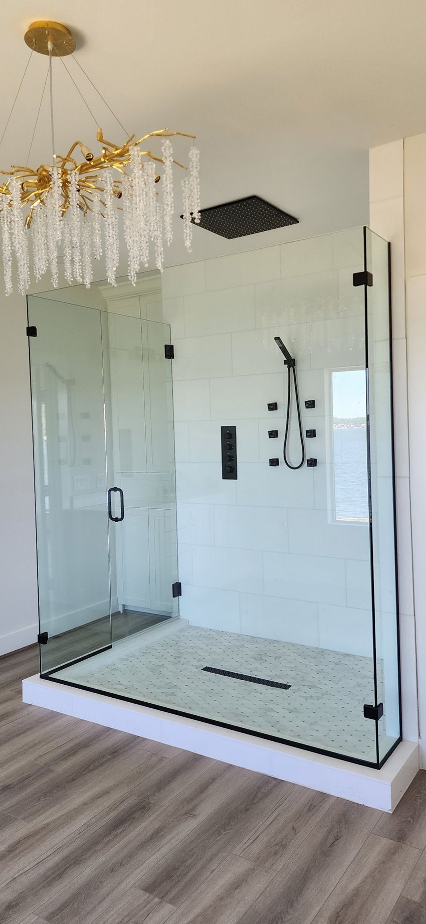 A modern walk-in glass shower with black hardware, a rainfall showerhead, and a chandelier above, set on light wood flooring.