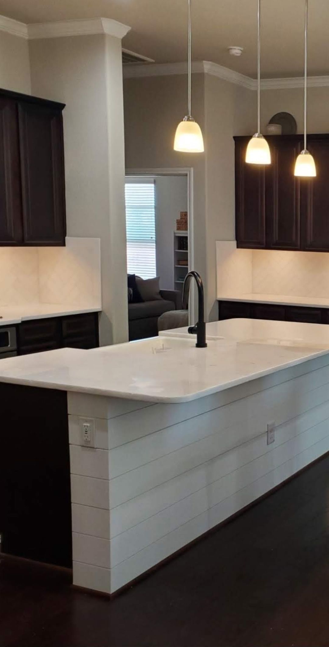 A kitchen island with a white countertop and white shiplap base, featuring a sink, dark cabinets, and three hanging lights.