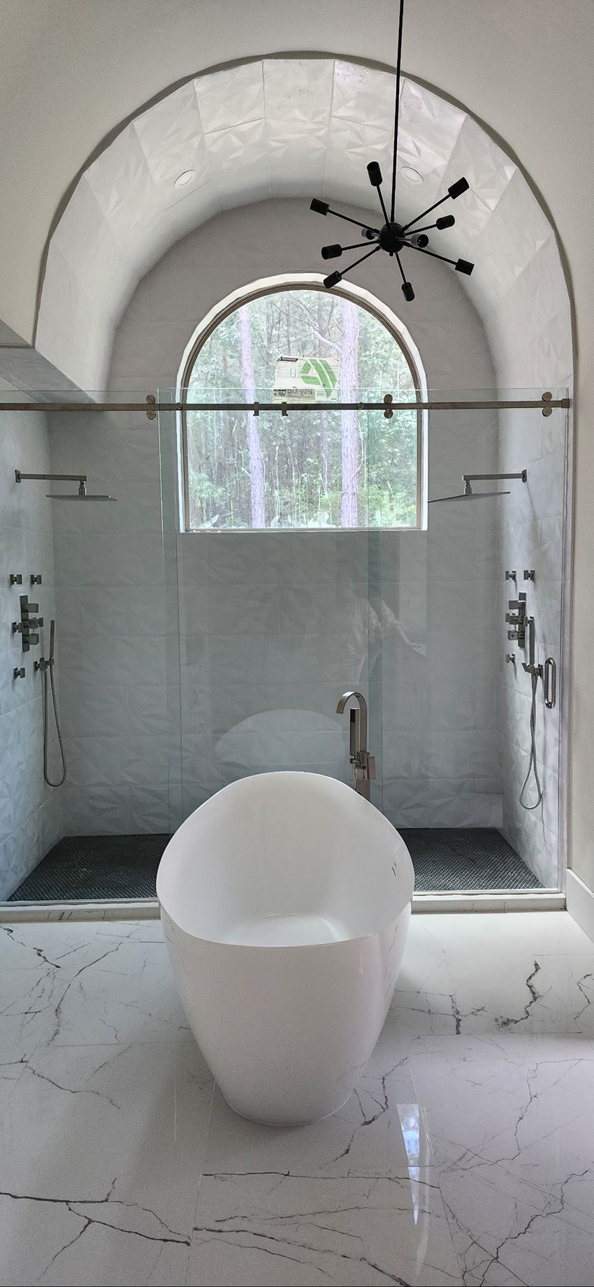 A modern bathroom featuring a white soaking tub, a double shower with glass doors, and a large arched window with a view.