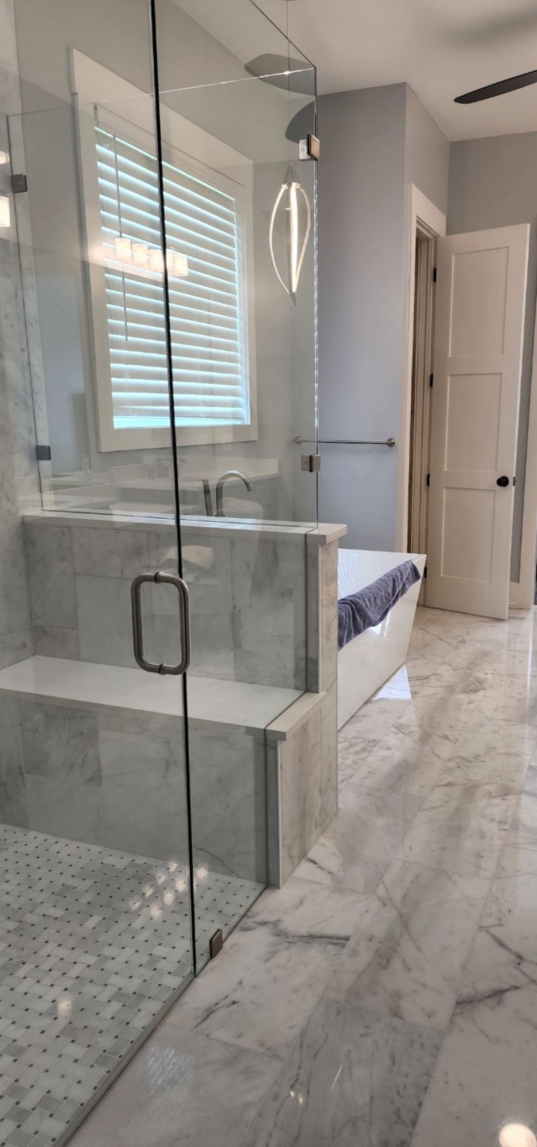 A modern bathroom featuring a glass-enclosed shower with marble tile, a built-in bench, and a bathtub under a window.