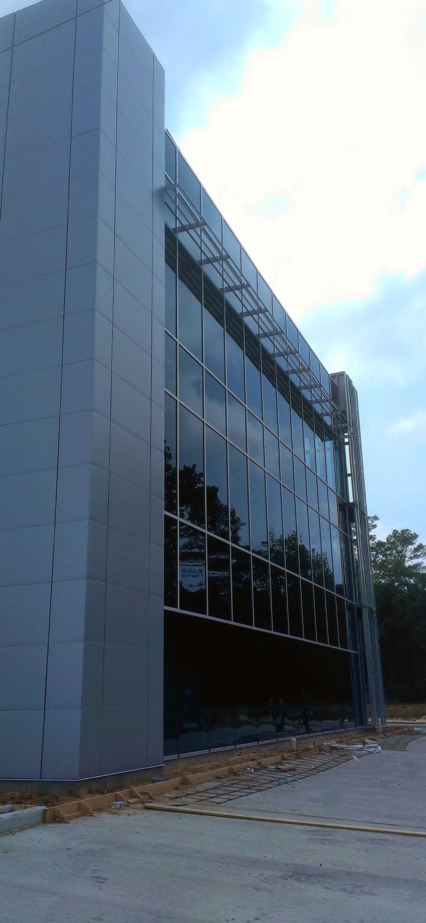 Low-angle view of a modern building facade with gray panels, a glass window wall, and exposed metal scaffolding.