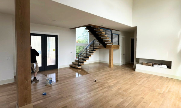 Bright empty living room with wood floors, staircase, and a person by the doorway.