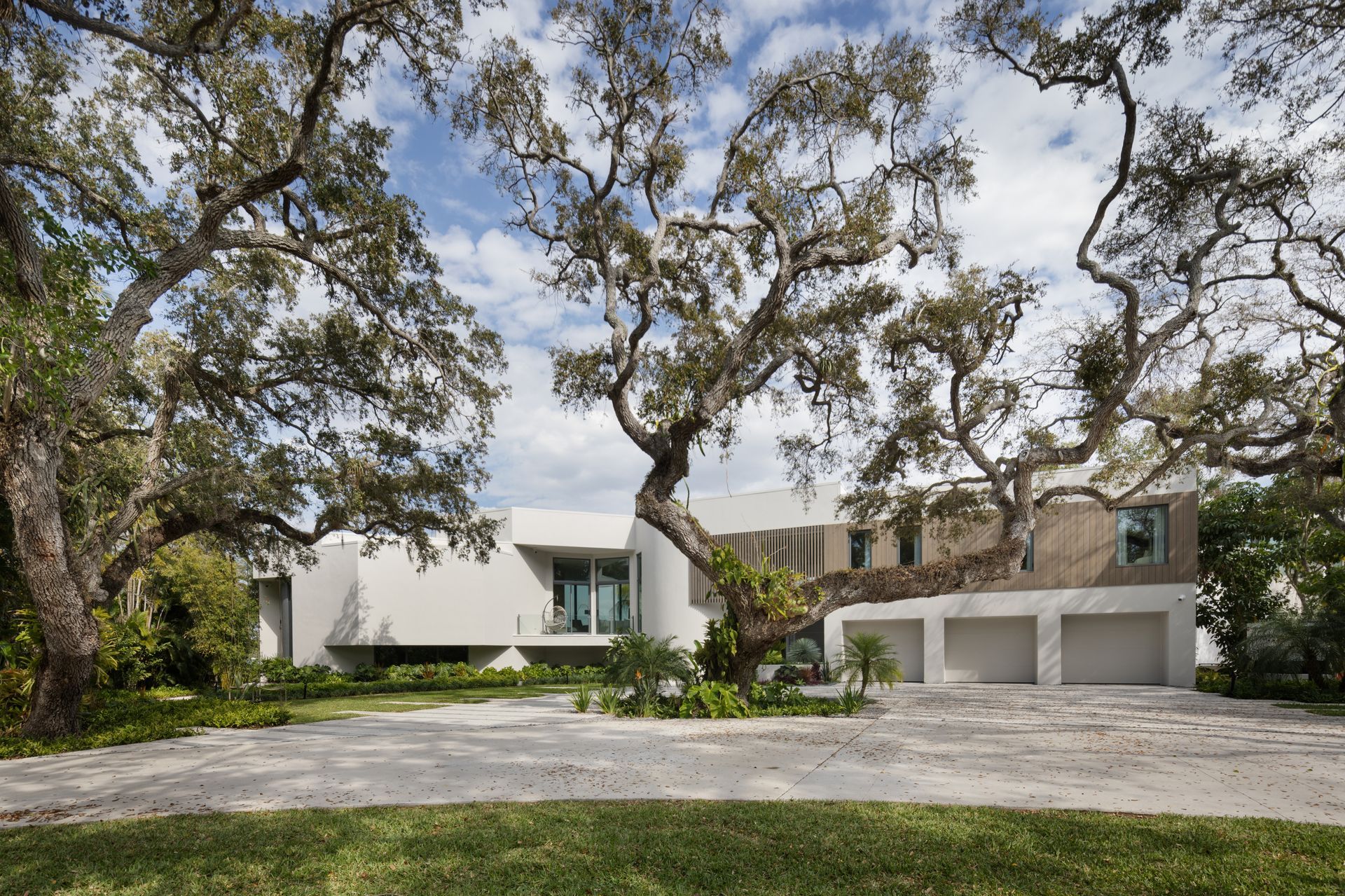 Modern white house with three-car garage and large trees. Concrete driveway. Blue sky with clouds.