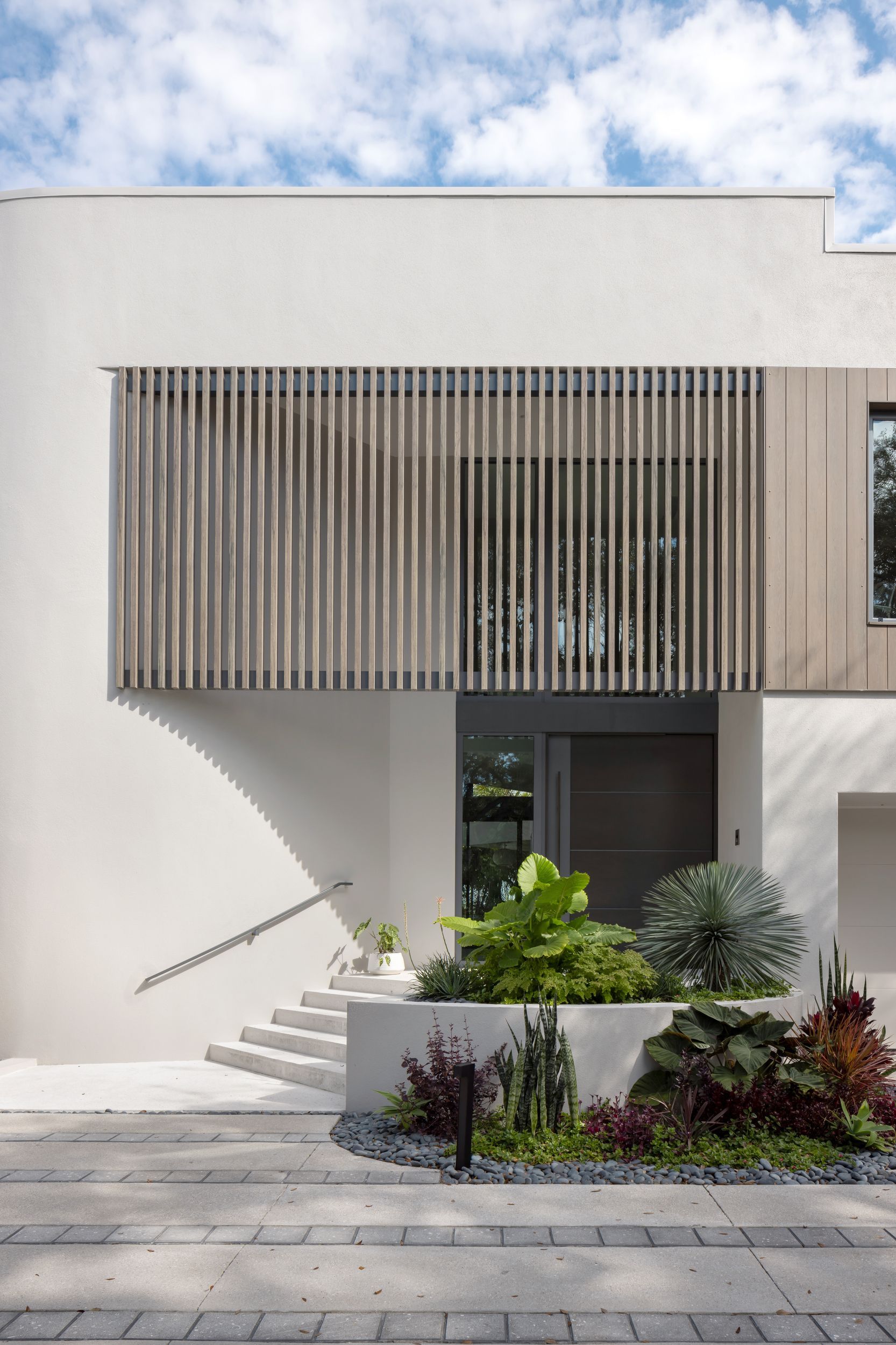 Modern white house entrance with wooden slats over windows, steps leading up, and a small garden.