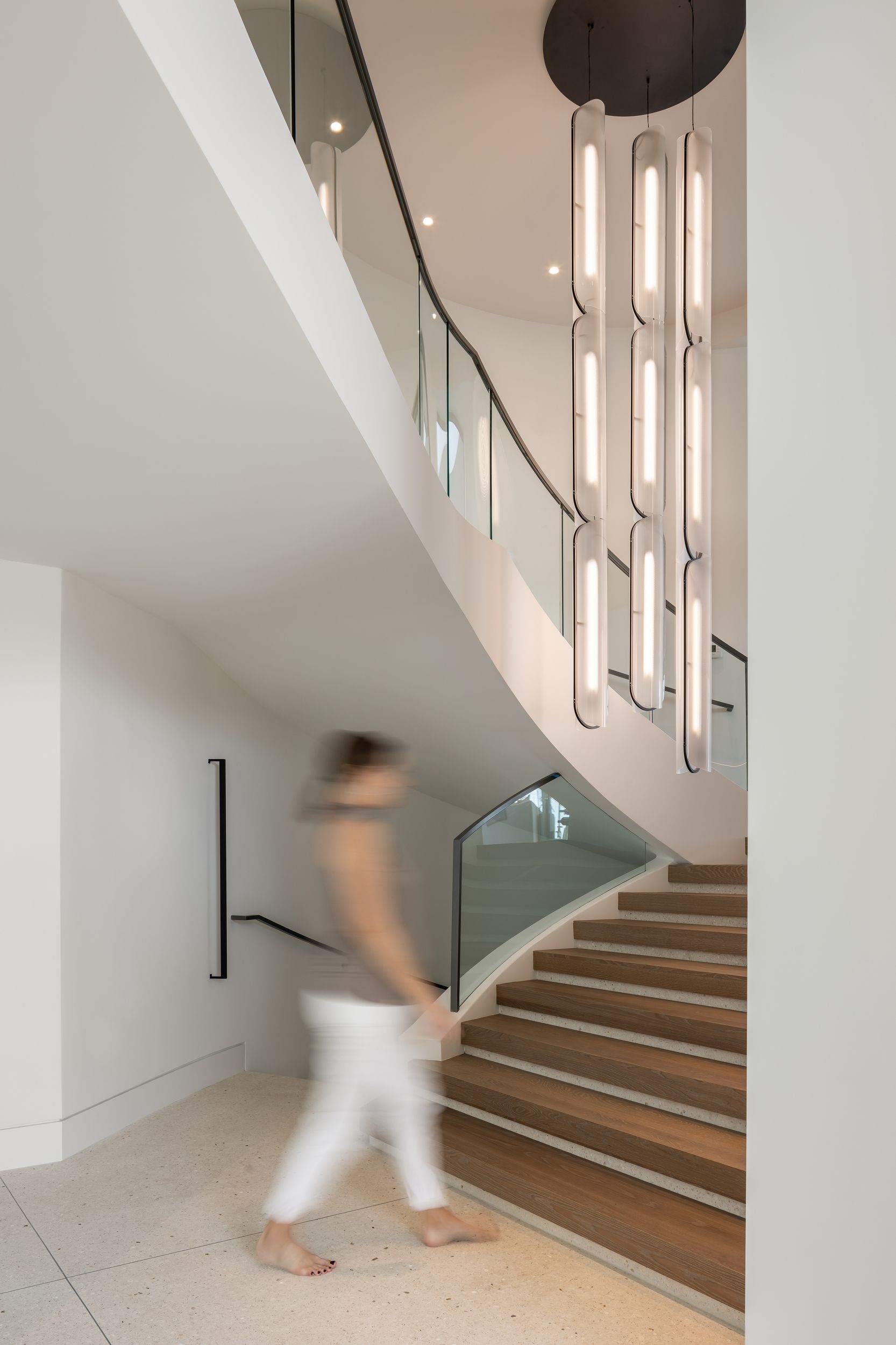 Person walking up a curved staircase with glass railing. Modern light fixture hangs overhead.