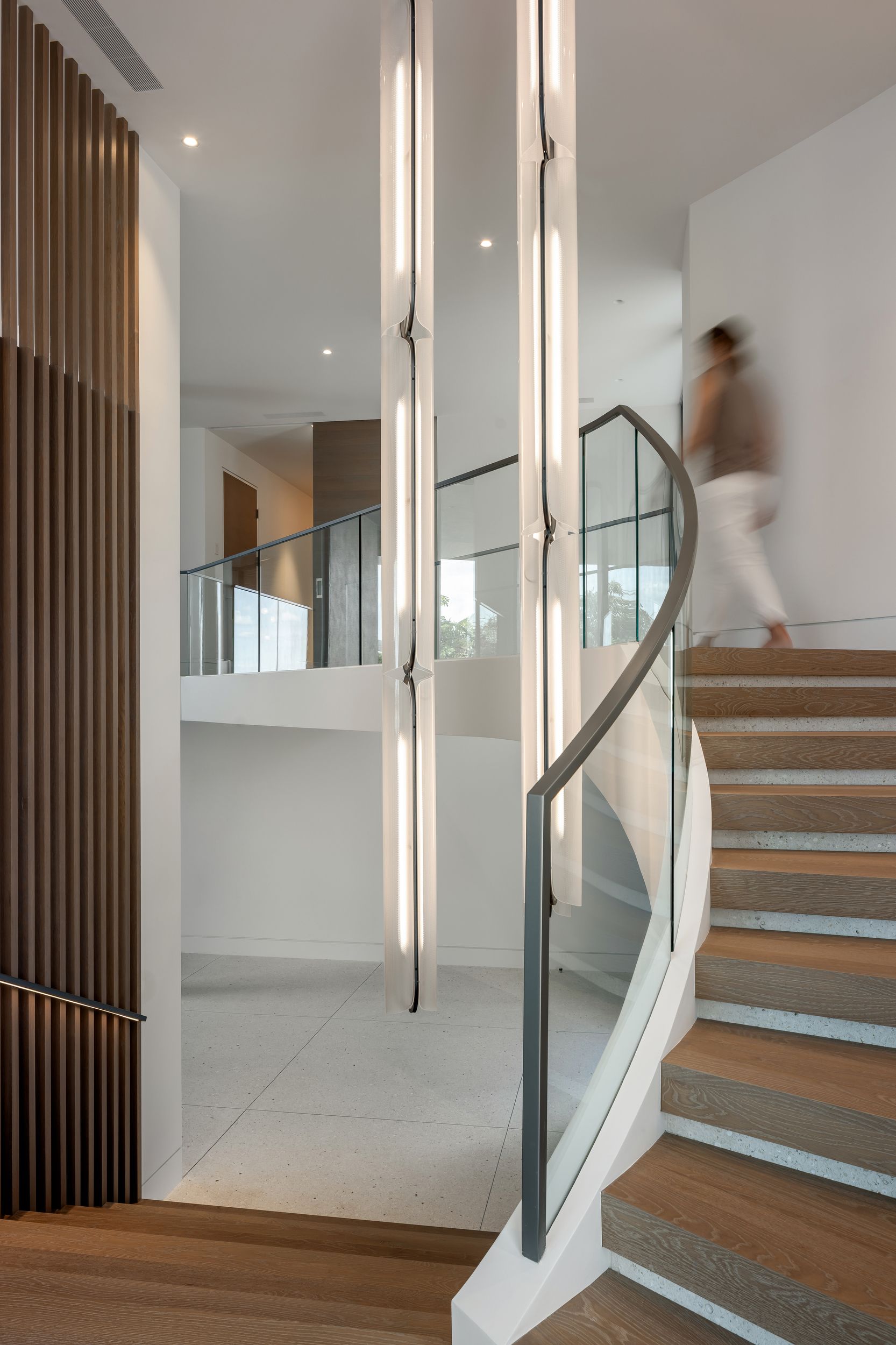 Modern stairwell with floating light fixtures, curved staircase, and person descending. Neutral tones, architectural design.