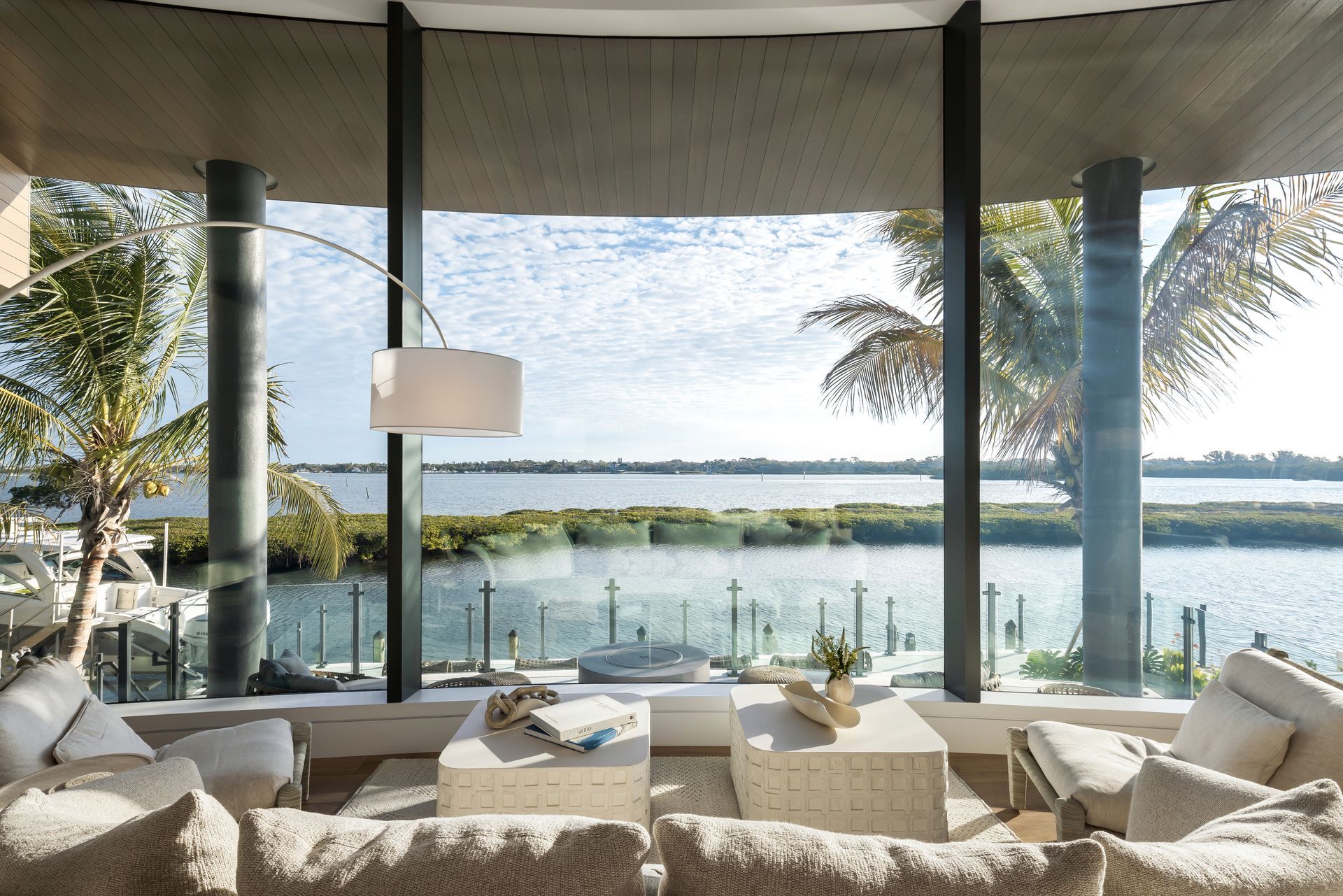 Lounge area with large windows overlooking a waterfront view, featuring neutral-toned furniture and a floor lamp.