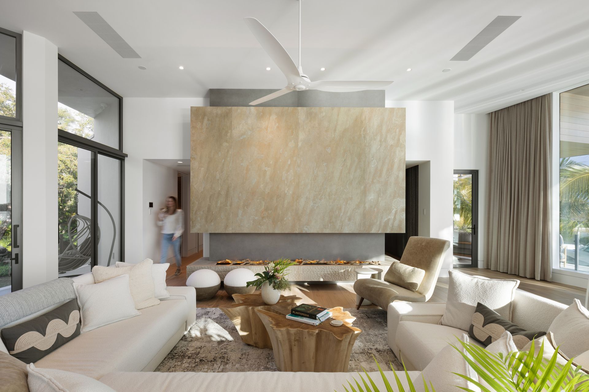 Modern living room with fireplace, white sofas, large windows, and a person in the doorway.