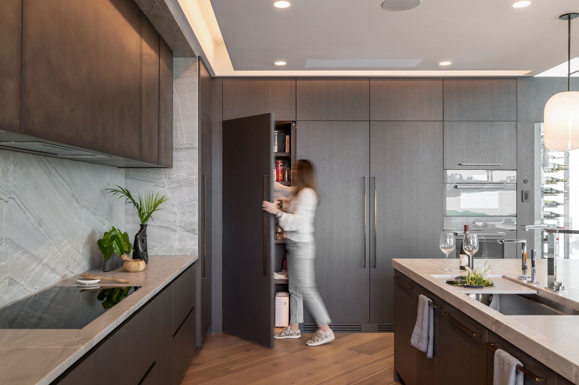Woman opening cabinet in modern kitchen with dark wood cabinetry and island.