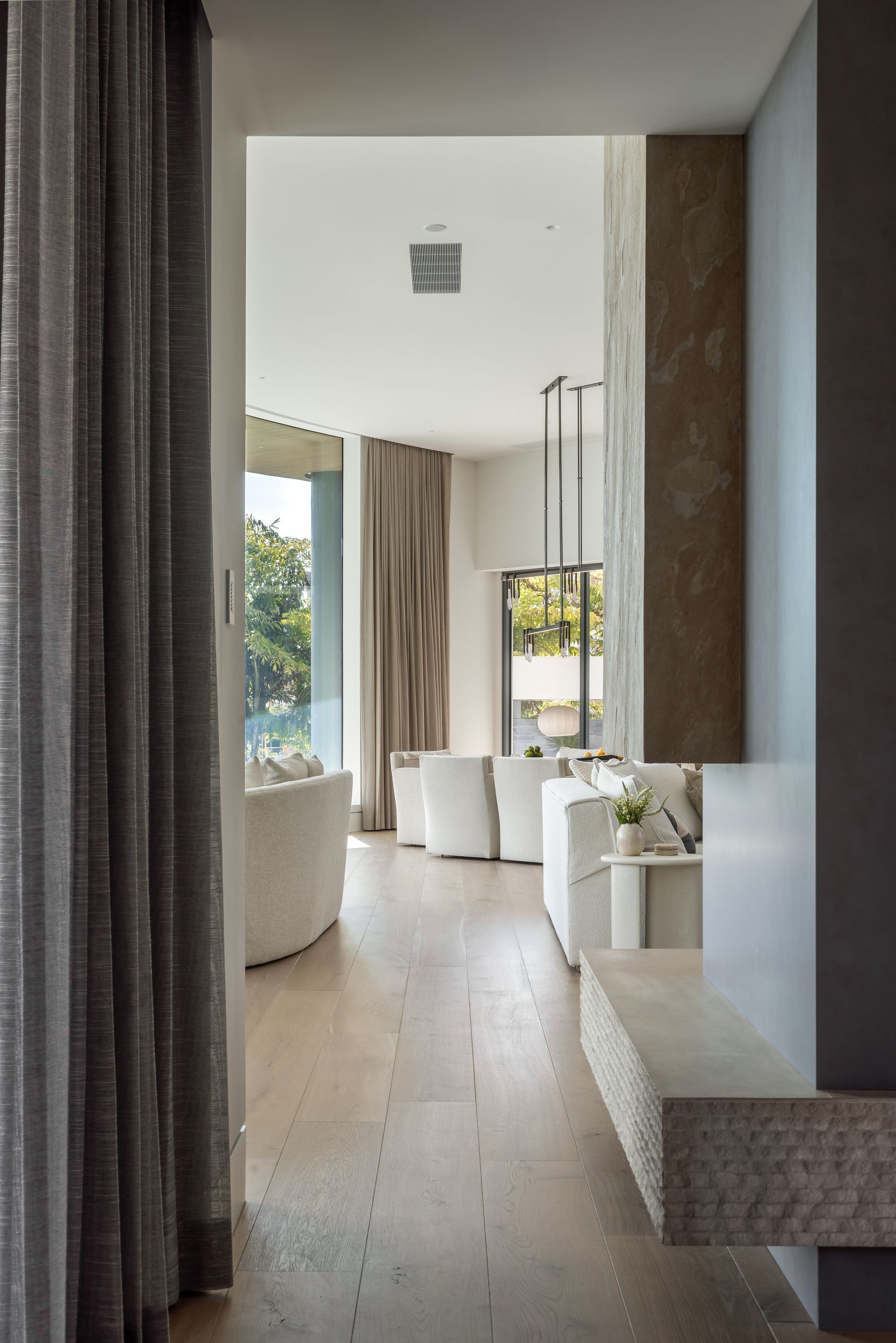 Hallway with light wood floors, a bench, and a view into a bright, modern interior with seating and natural light.
