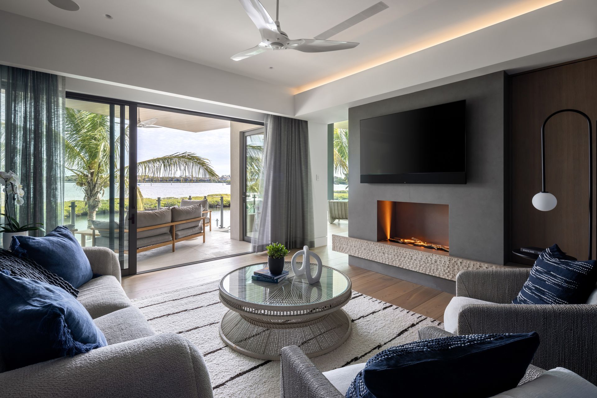 Living room with open doors to a patio overlooking water.  Gray sofa, rug, and fireplace. Large TV above the fireplace.