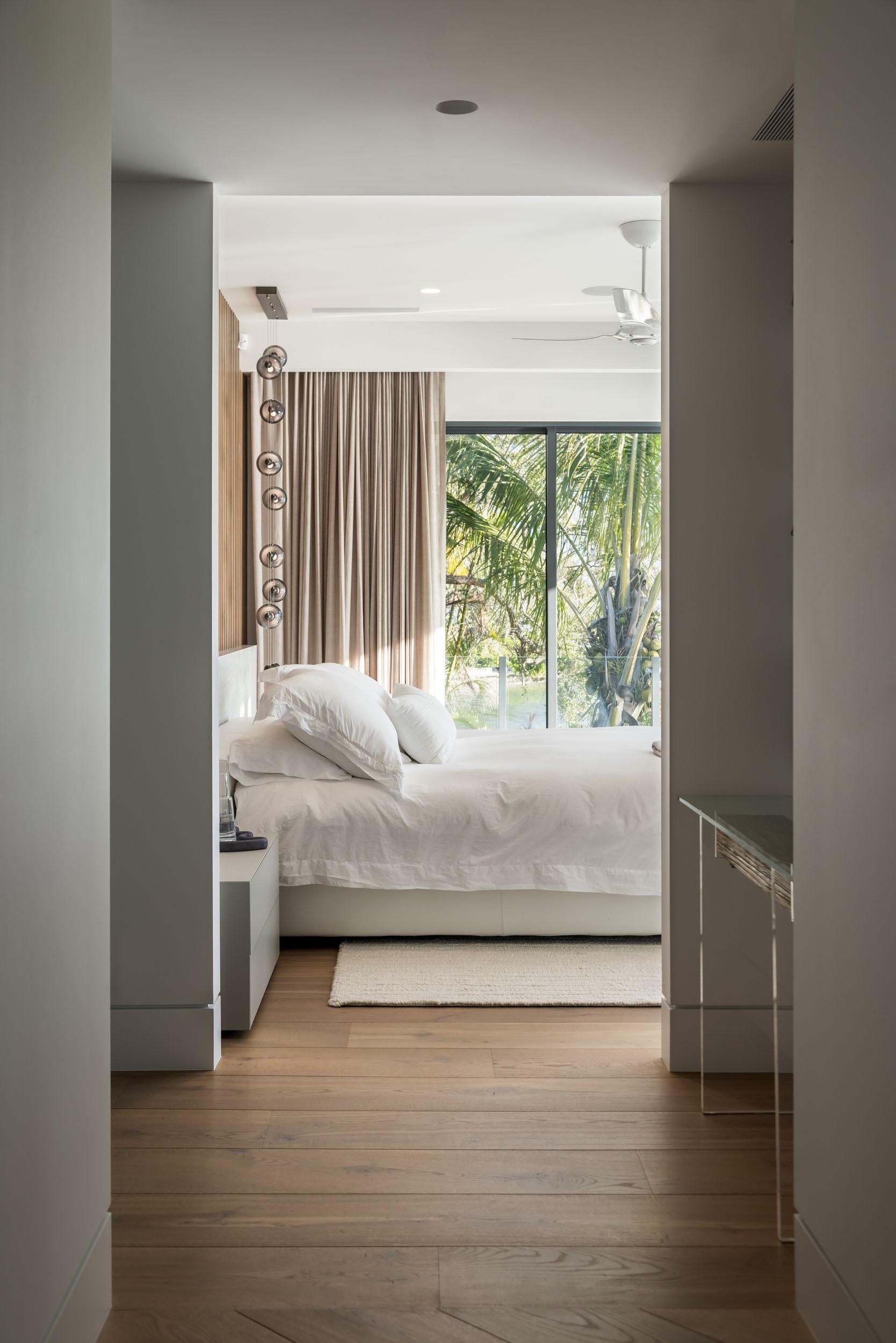 Bedroom interior: white bedding, tan curtains, wood floor, view to a window overlooking greenery.