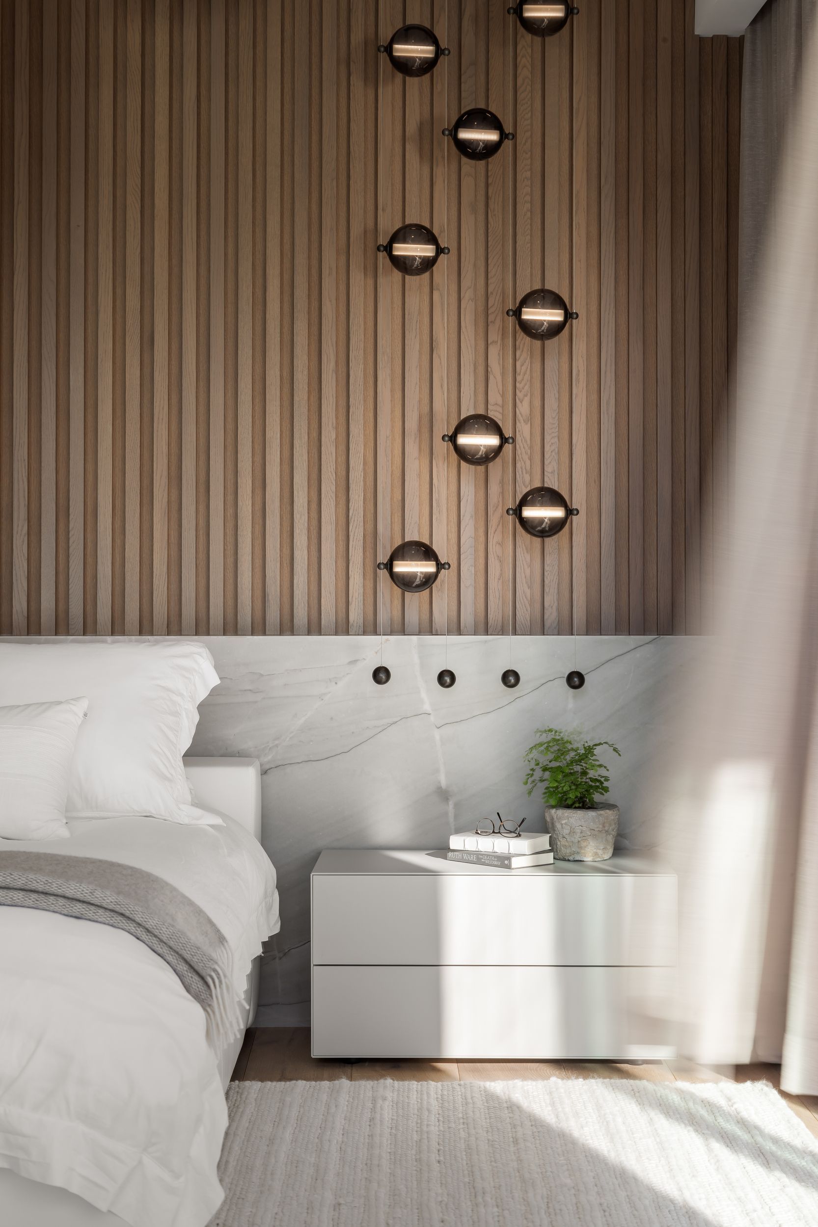 Bedroom with a white bed, wood-paneled wall, modern sconces, white nightstand, and potted plant.