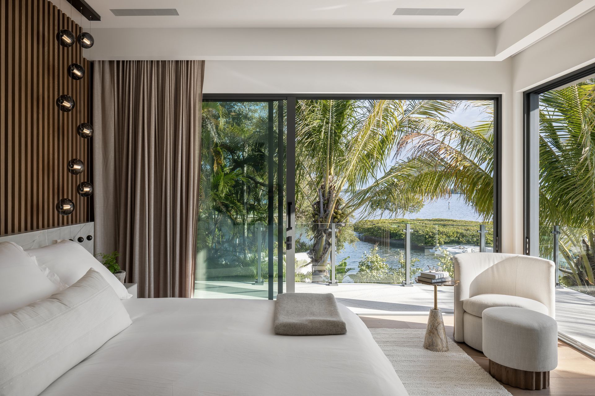 Bedroom with king bed, sliding glass doors overlooking water and trees, and a beige armchair.