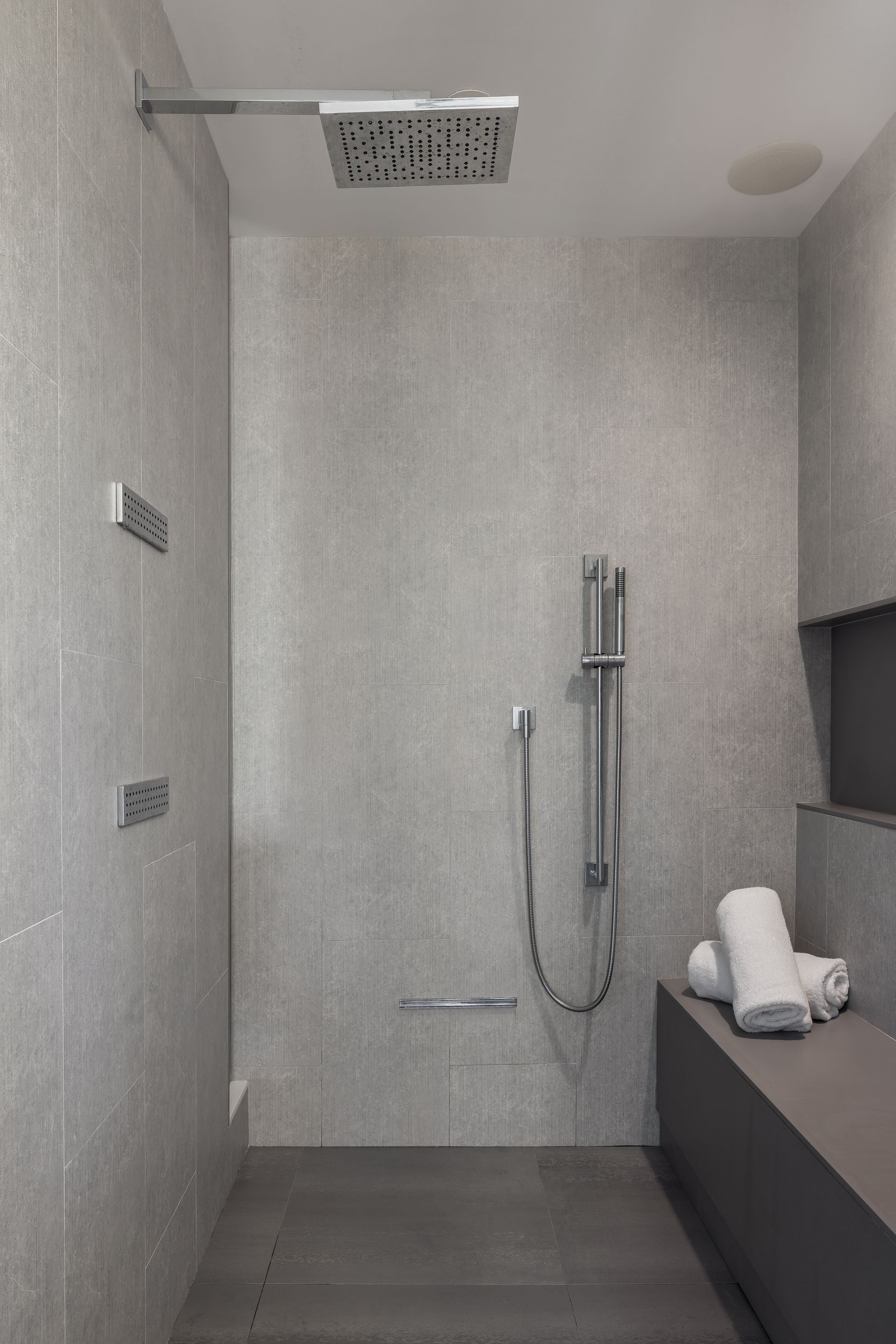 Modern gray tiled shower with overhead showerhead, handheld shower, bench, and towels.