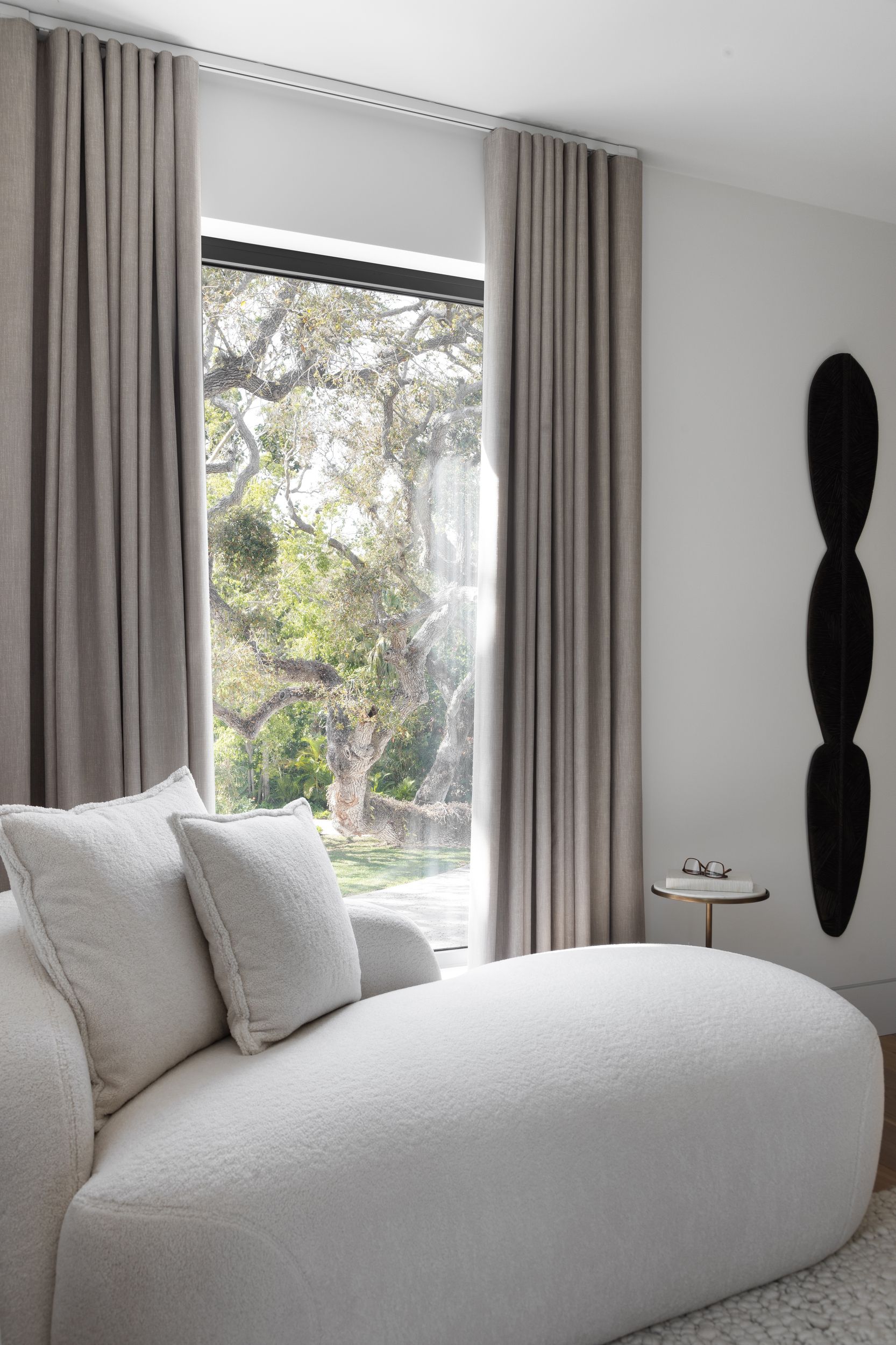 Cozy white sofa by window with neutral curtains, a minimalist art piece, and a small table.