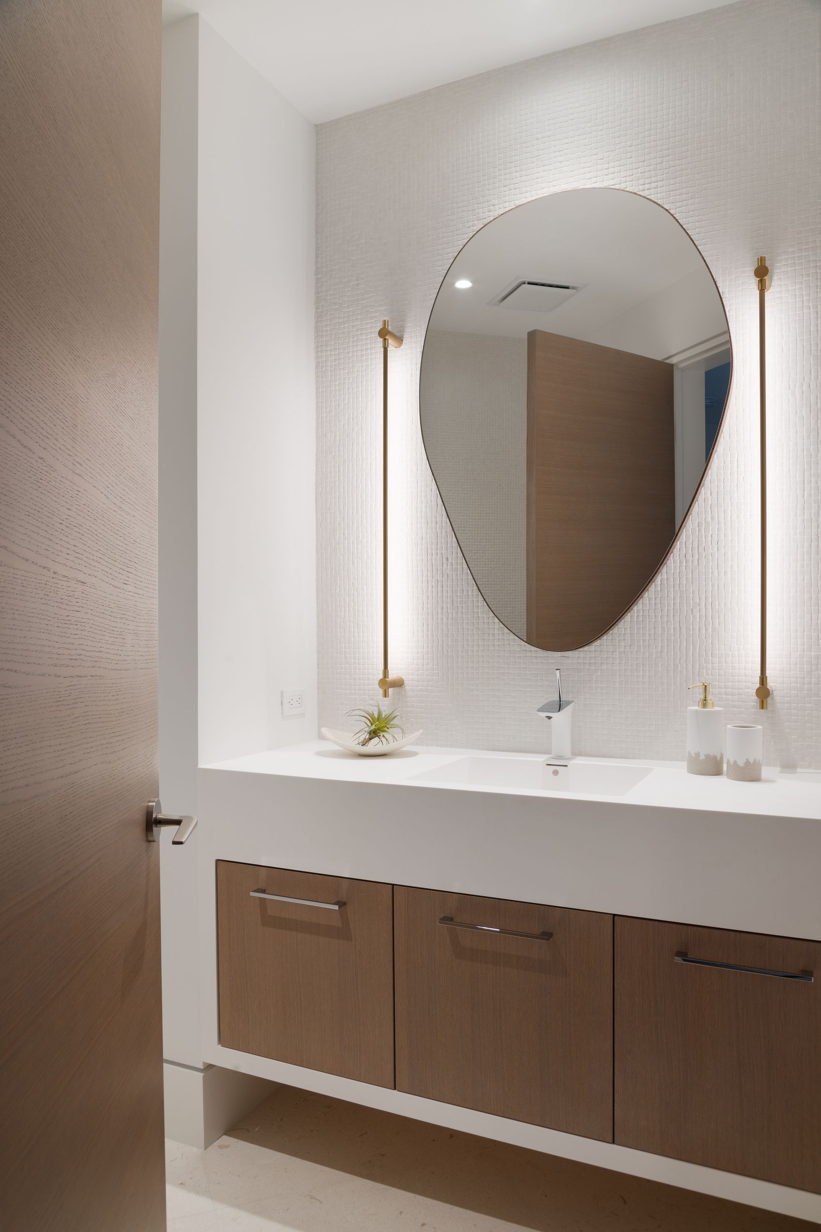 Modern bathroom with oval mirror, wood cabinets, and vertical sconces. White and light wood tones.