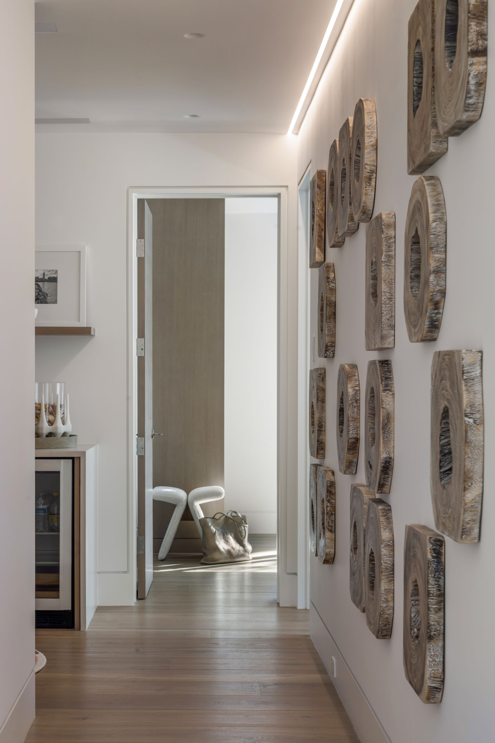 Narrow hallway with wooden floor, white walls, and abstract stone art. A doorway leads to a sunlit space.