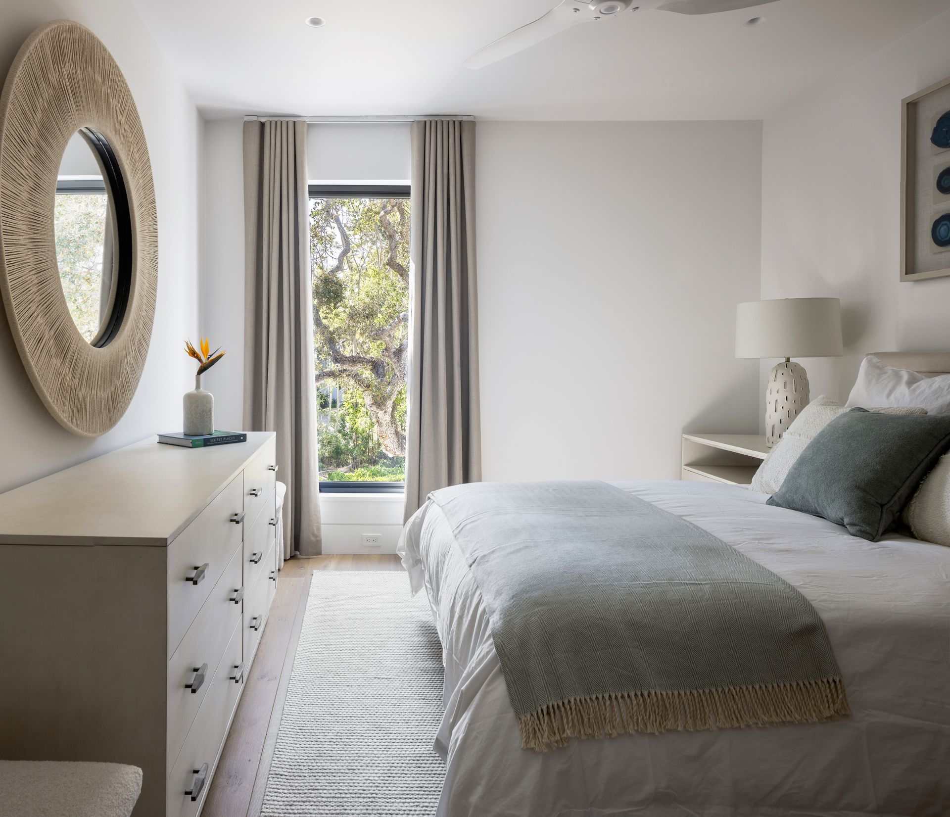 Bedroom with white walls, dresser, bed, mirror. Window shows trees, light gray curtains.