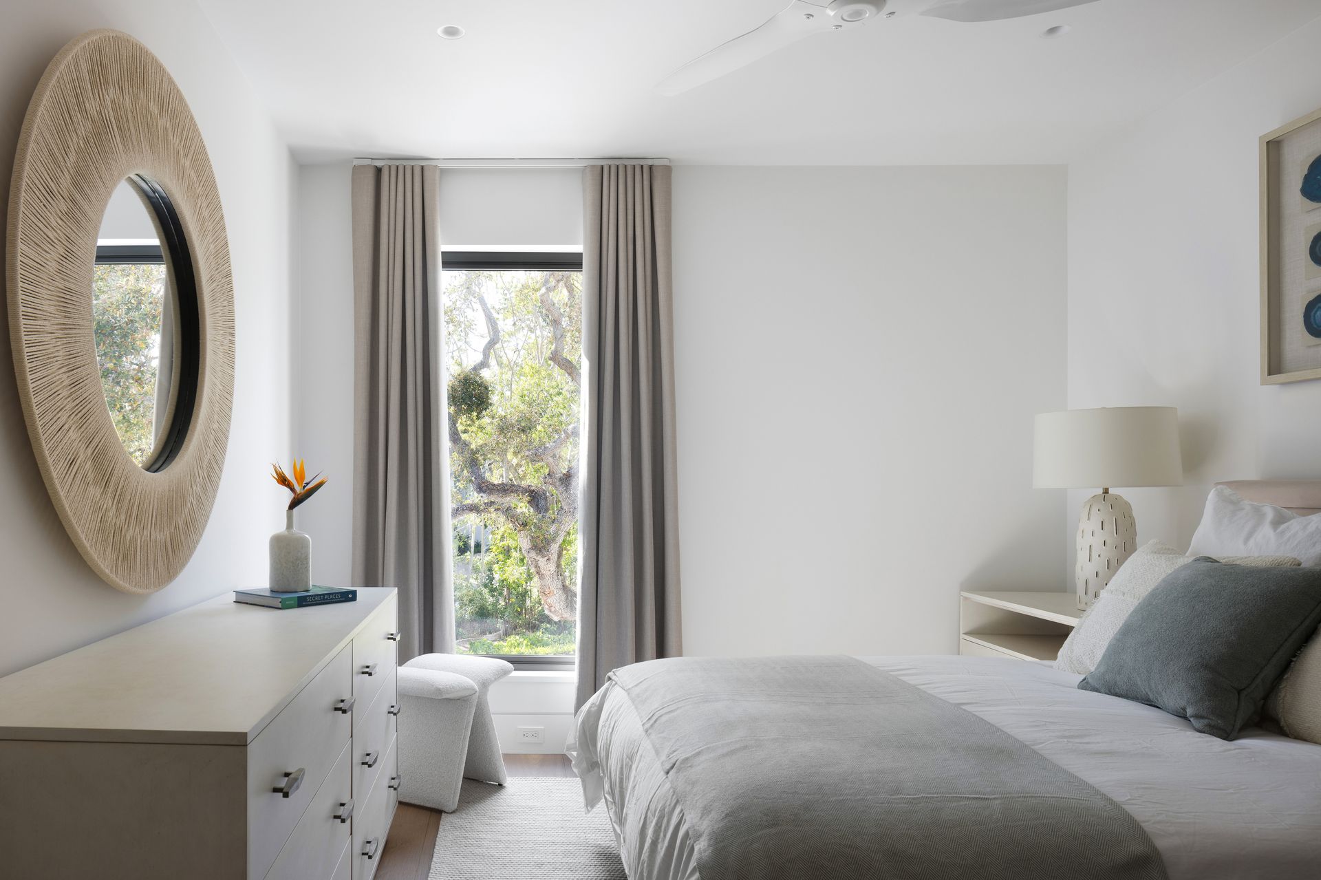 Bedroom with white walls, large mirror, bed with light gray blanket, and window with curtains.
